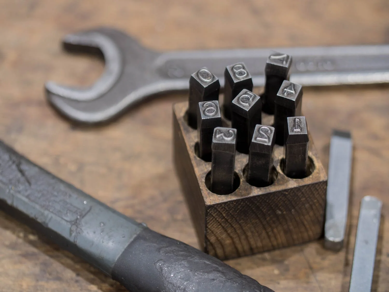 A wrench, a wooden block with numbered black metal stamps, and two metal rods on a wooden surface.
