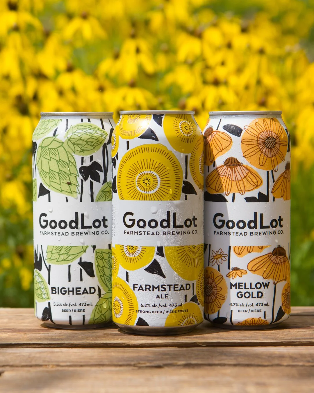 Three cans of GoodLot Farmstead Brewing Co. beer with floral and leaf designs, placed on a wooden surface against a yellow flower field background.