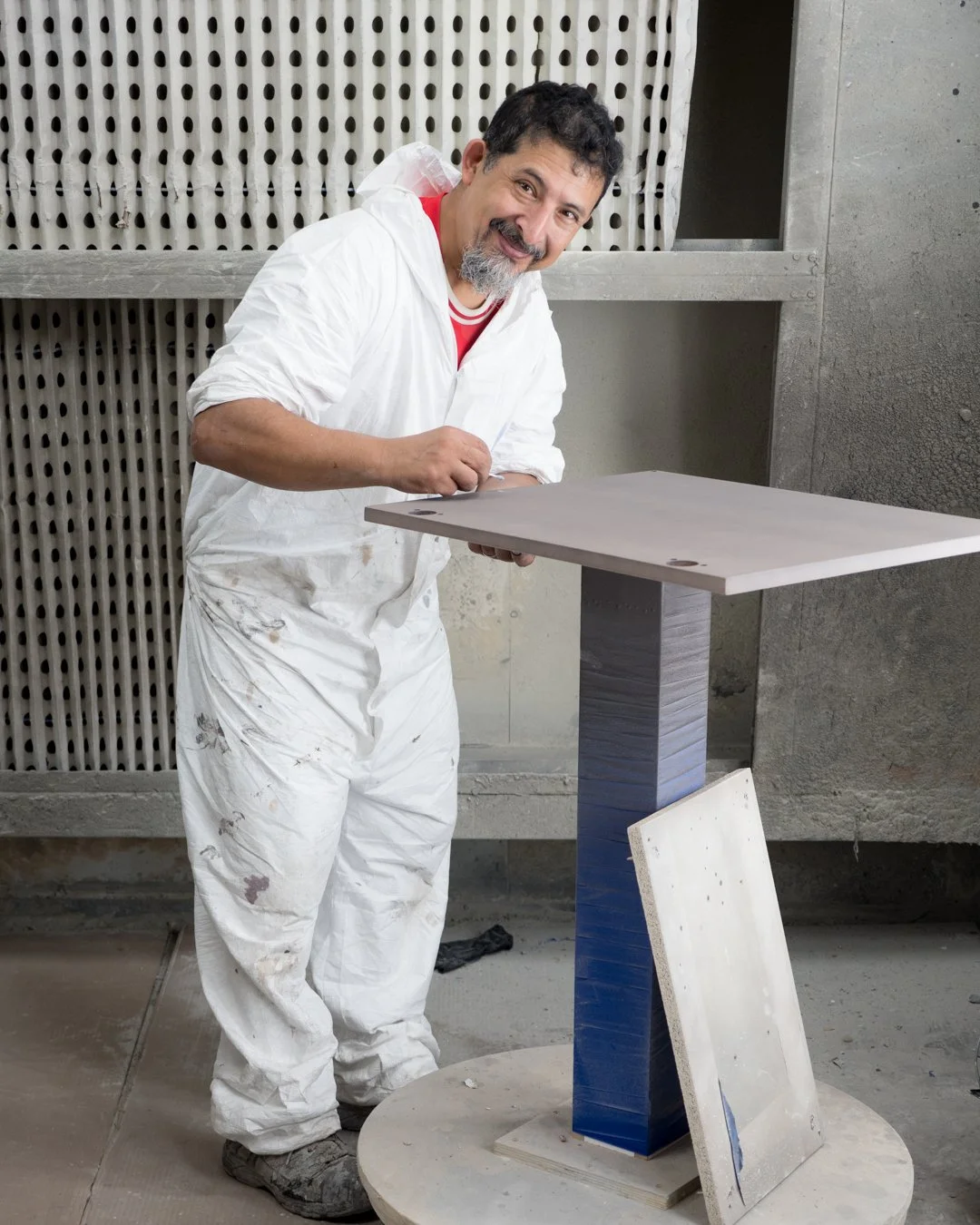 A man with a beard and curly hair, wearing white work clothes, is smiling while working on a table in a factory setting. The background has a concrete wall and a perforated metal panel.