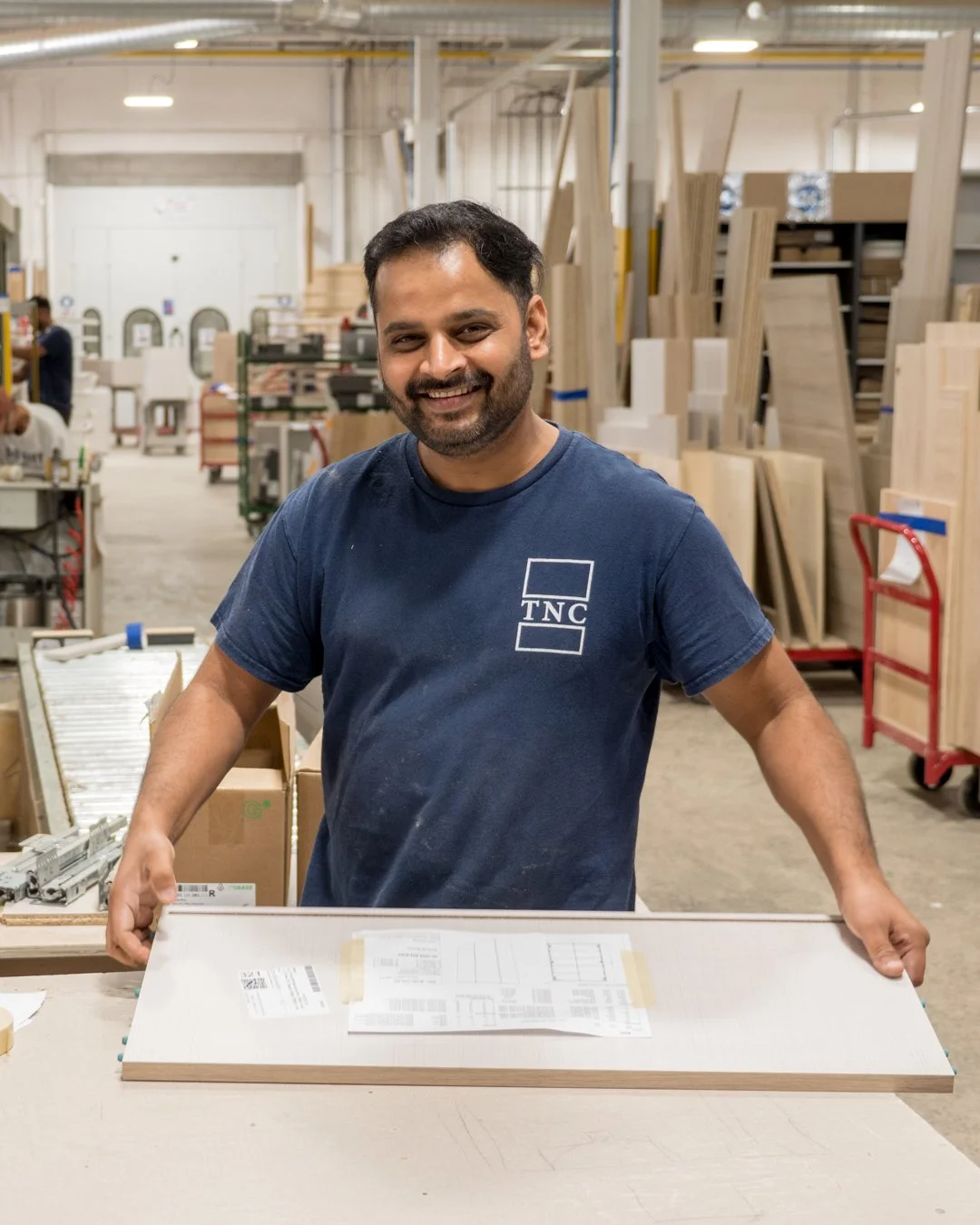A smiling man with dark hair and beard in a woodworking shop, holding a wooden board with plans on it. The shop has shelves of wood pieces and equipment in the background.