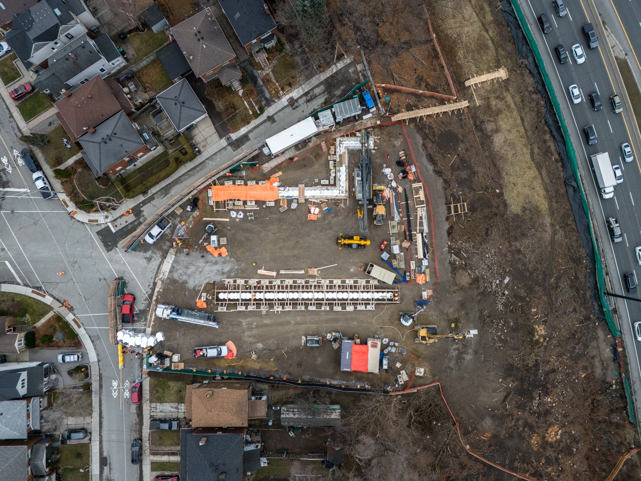 Aerial view of a construction site adjacent to a residential neighborhood and a highway, showing ongoing building work, construction equipment, materials, and surrounding houses.