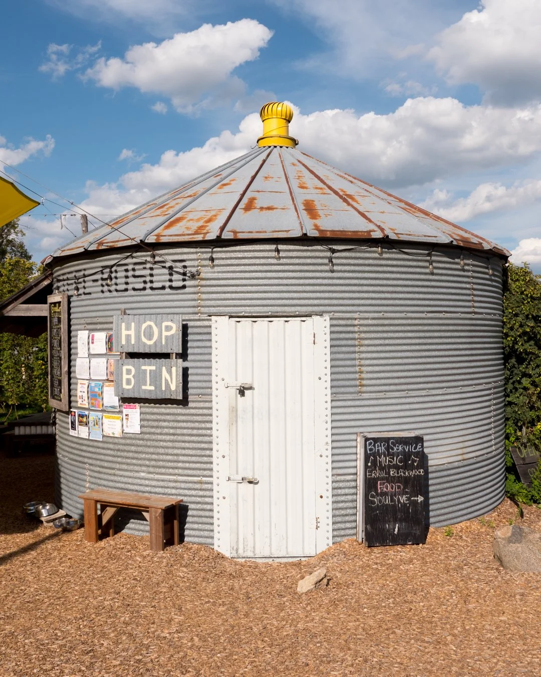 A round, metal grain silo with a rusted roof, used as a hop bin for a brewery. Signs advertise hop bin, bar service, music, and food. There's a small wooden bench and food bowls in front of it.
