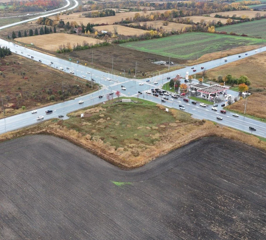 Aerial view of a rural intersection with a traffic signal, a convenience store, and several vehicles on the roads, surrounded by farmland and fields.