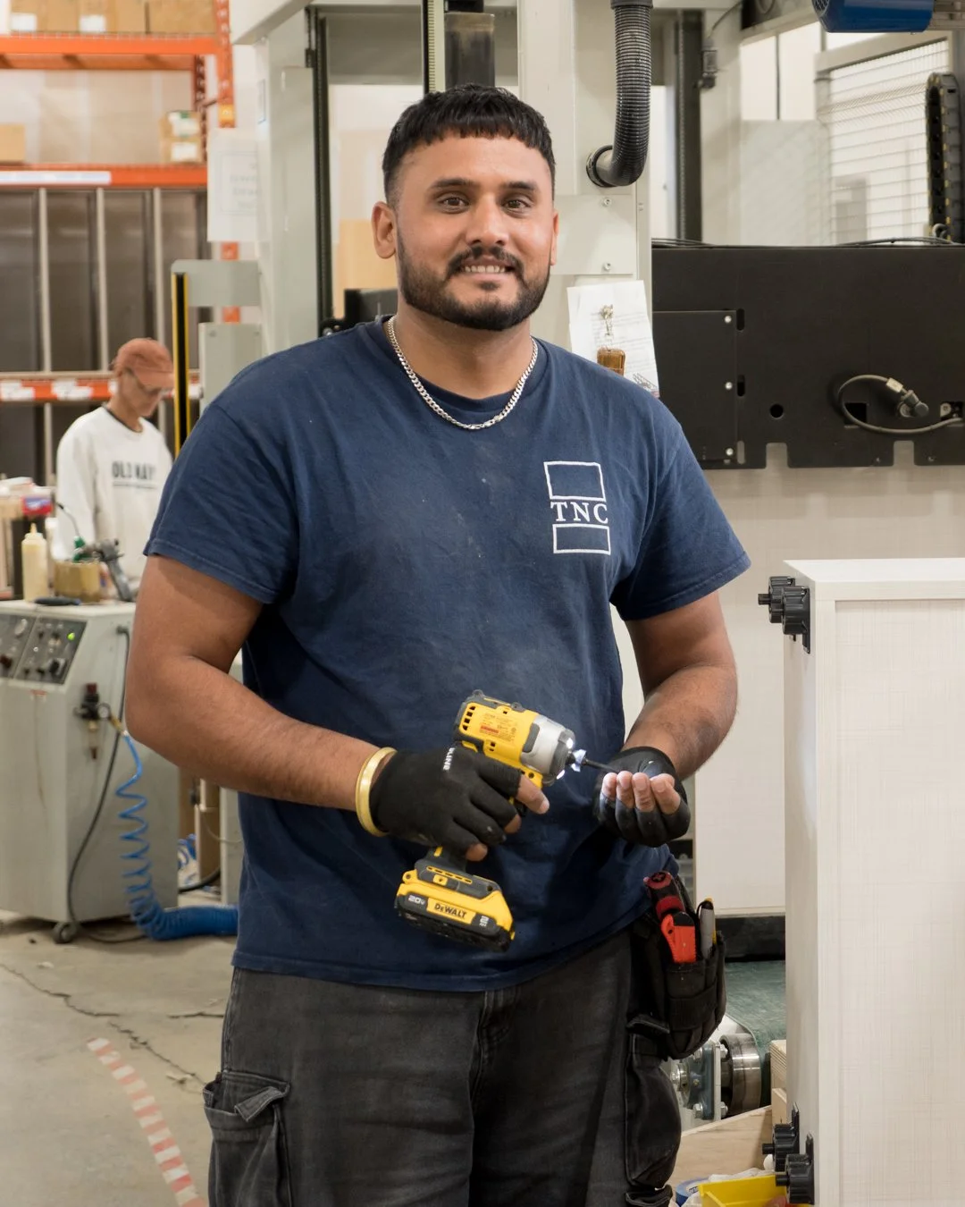 A man working with a cordless drill in a factory setting, wearing a blue TNC t-shirt, gloves, and a tool pouch.