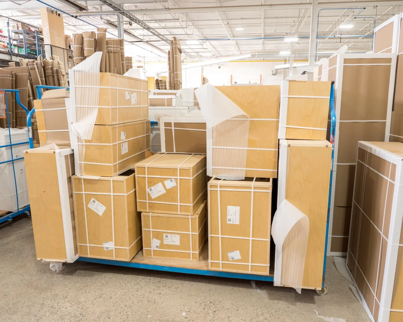 Various flat-packed wooden furniture and cabinet boxes stored on a blue cart in a warehouse aisle.