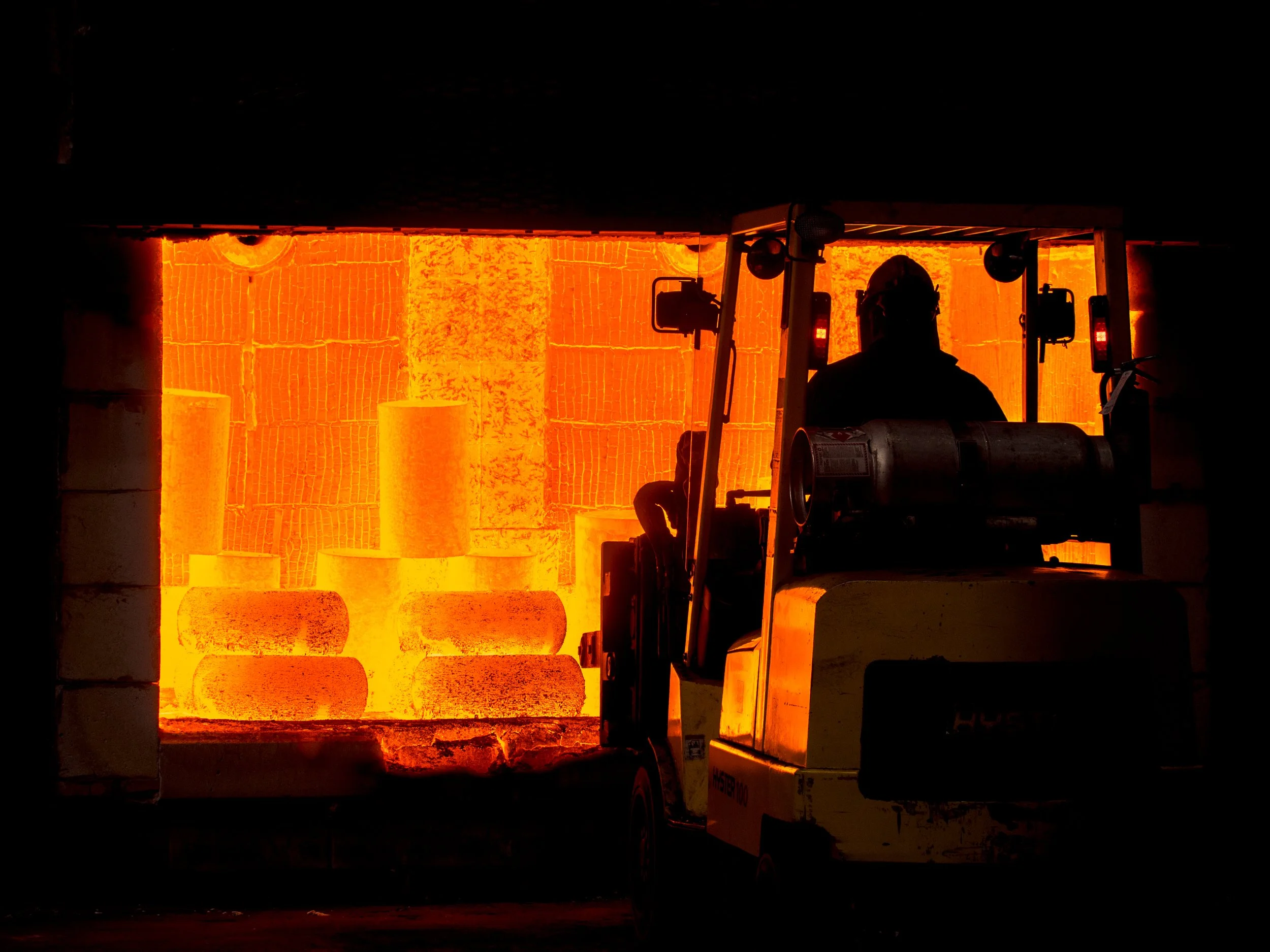 A worker operating a forklift inside a foundry with a glowing, fiery furnace in the background, pouring molten metal into molds.