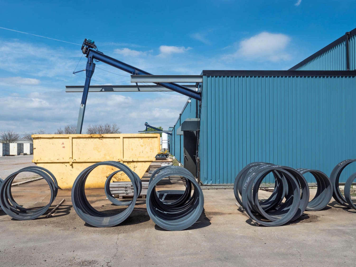 Industrial equipment outdoors with large blue storage building, metal coils in foreground, crane extending from building, and a large yellow container.