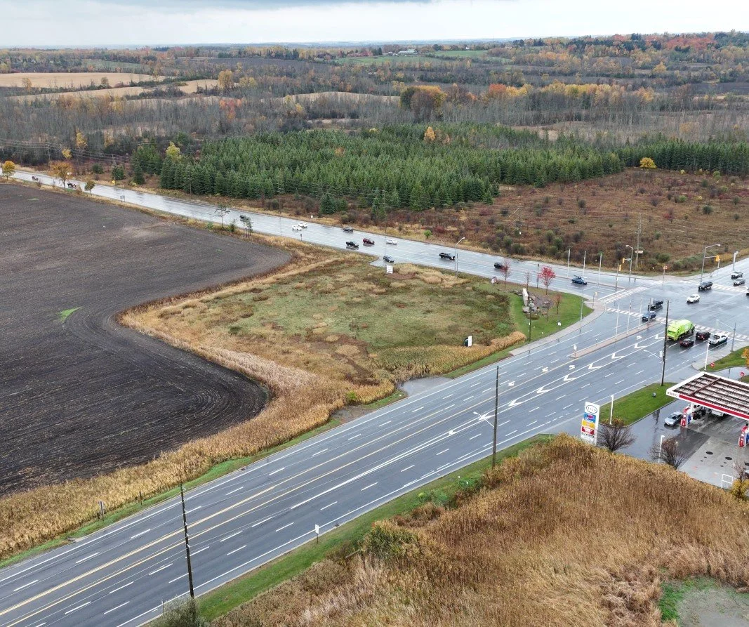 Aerial view of a rural intersection with a gas station, nearby farmland, and forested areas in the background, featuring wet roads and vehicles.