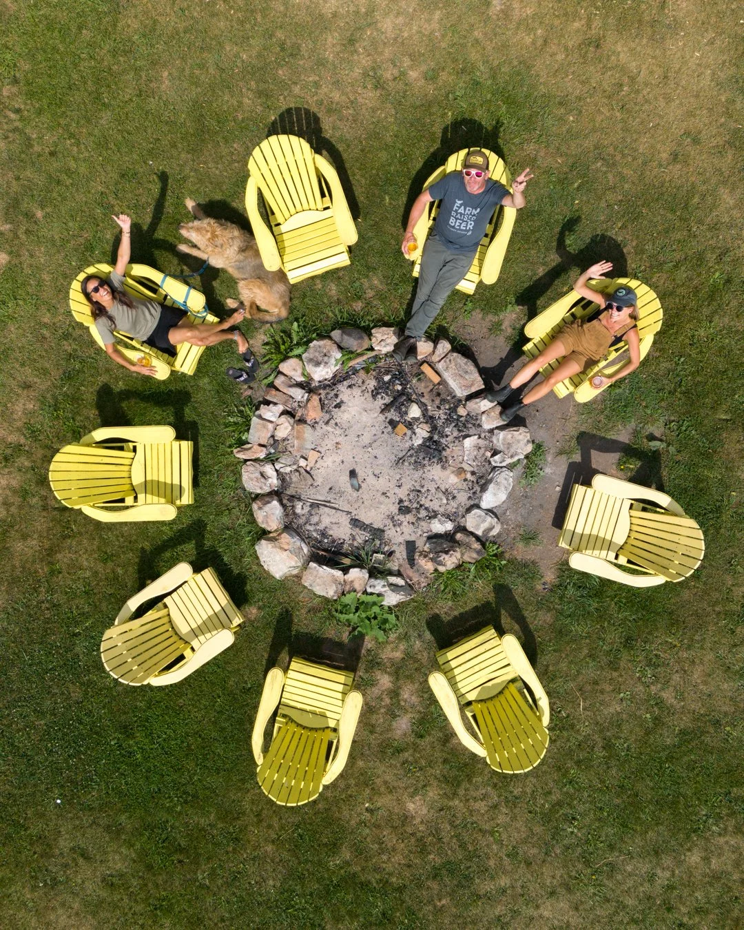 Aerial view of four people sitting on yellow Adirondack chairs arranged in a circle around a fire pit, with two dogs nearby on grass.