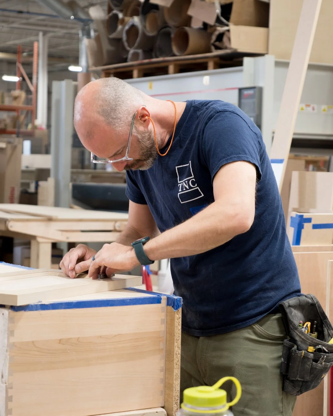 A man working on woodworking in a workshop, wearing safety glasses and a navy blue TNC t-shirt, focused on assembling or shaping wood pieces, with various tools and wood pieces in the background.