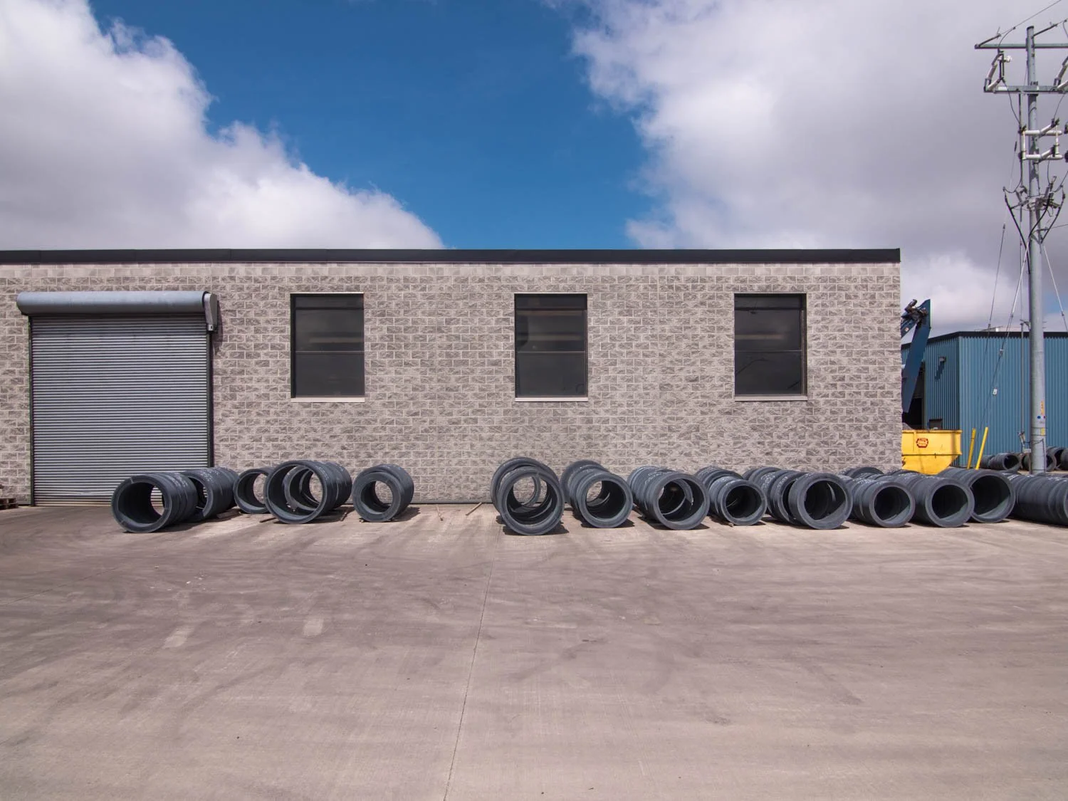 Industrial building with three windows, a roller shutter door, and large black pipes lined up outside on a concrete surface under a partly cloudy sky.
