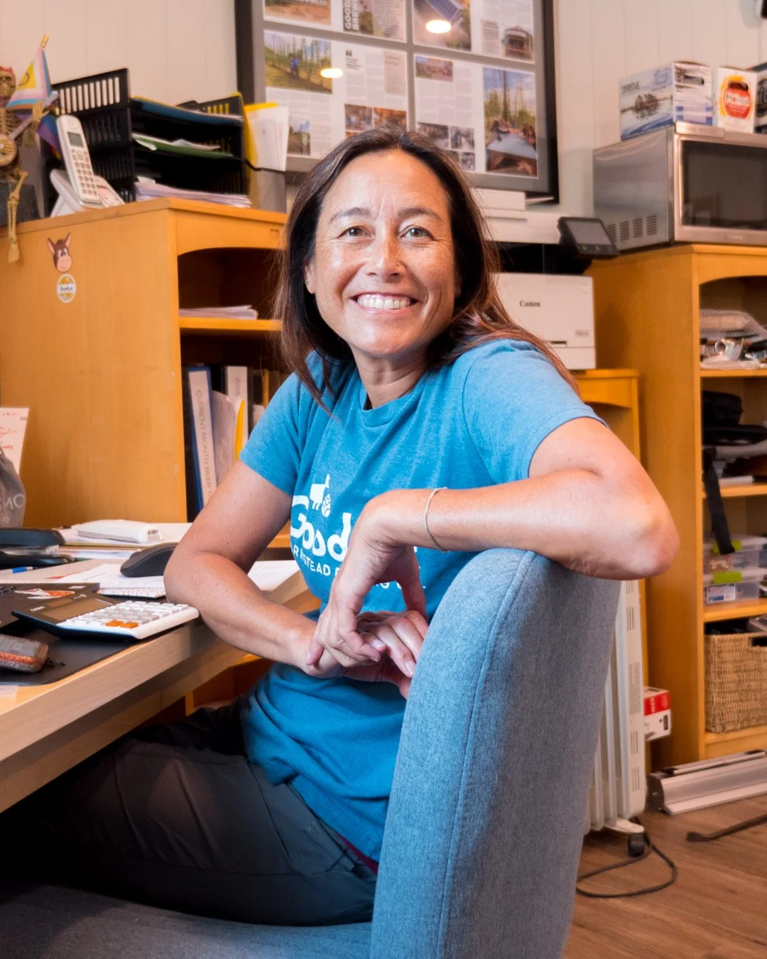 A woman smiling, sitting at a cluttered desk in an office, surrounded by shelves filled with papers and office equipment.