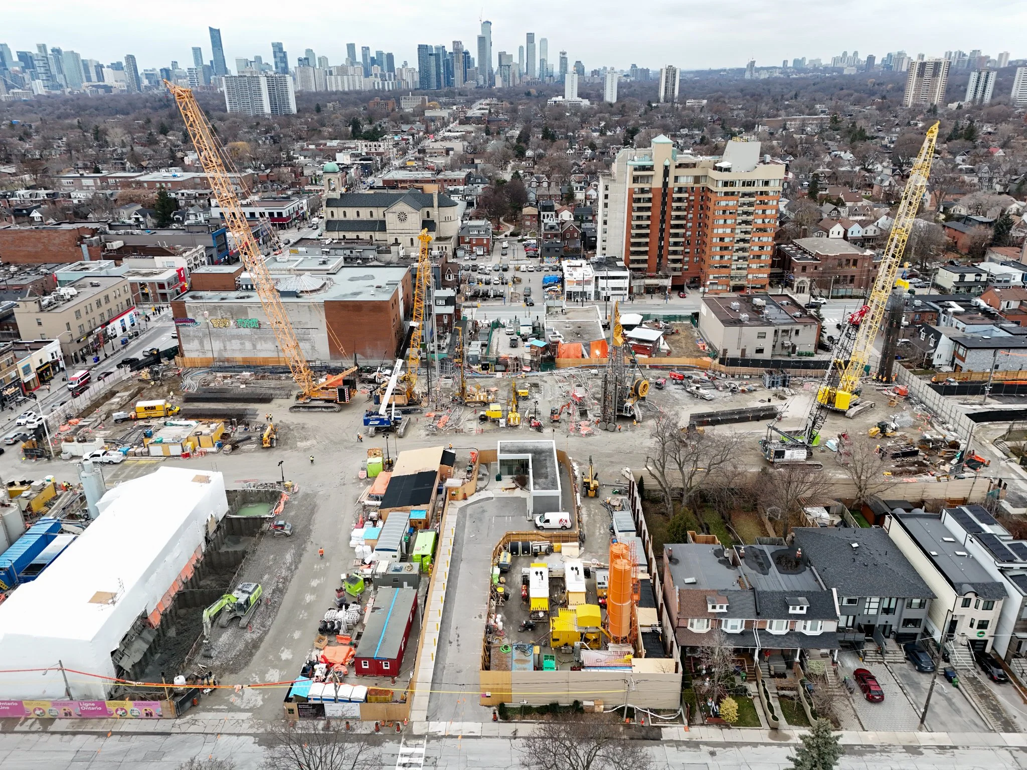 Aerial view of a city under construction with multiple cranes working on a construction site, surrounded by residential and commercial buildings, with a downtown skyline in the distance.