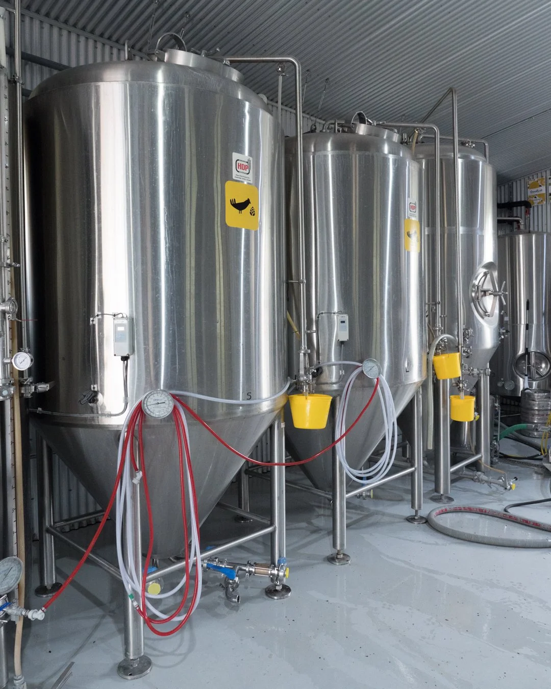 Several large stainless steel fermentation tanks with yellow and red tubing attached, located in a brewery or winery facility.