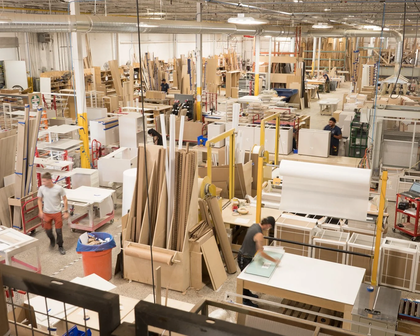 A busy woodworking workshop with workers surrounded by wood panels, machinery, and workstations.