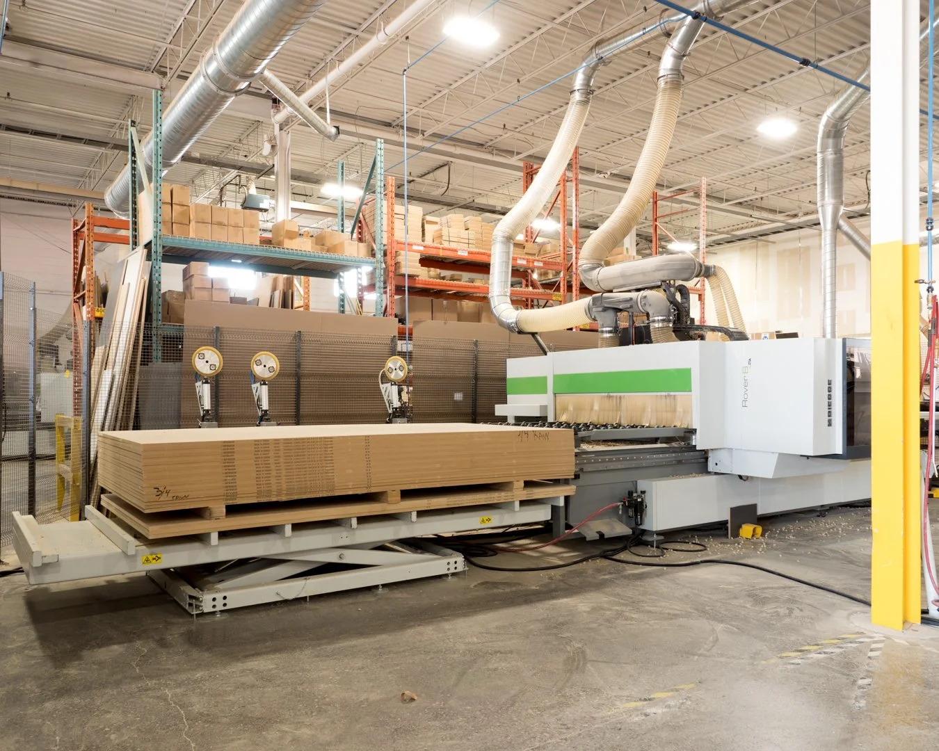 Woodworking factory with CNC machine cutting plywood sheets, shelves with boxes, and industrial ventilation pipes.