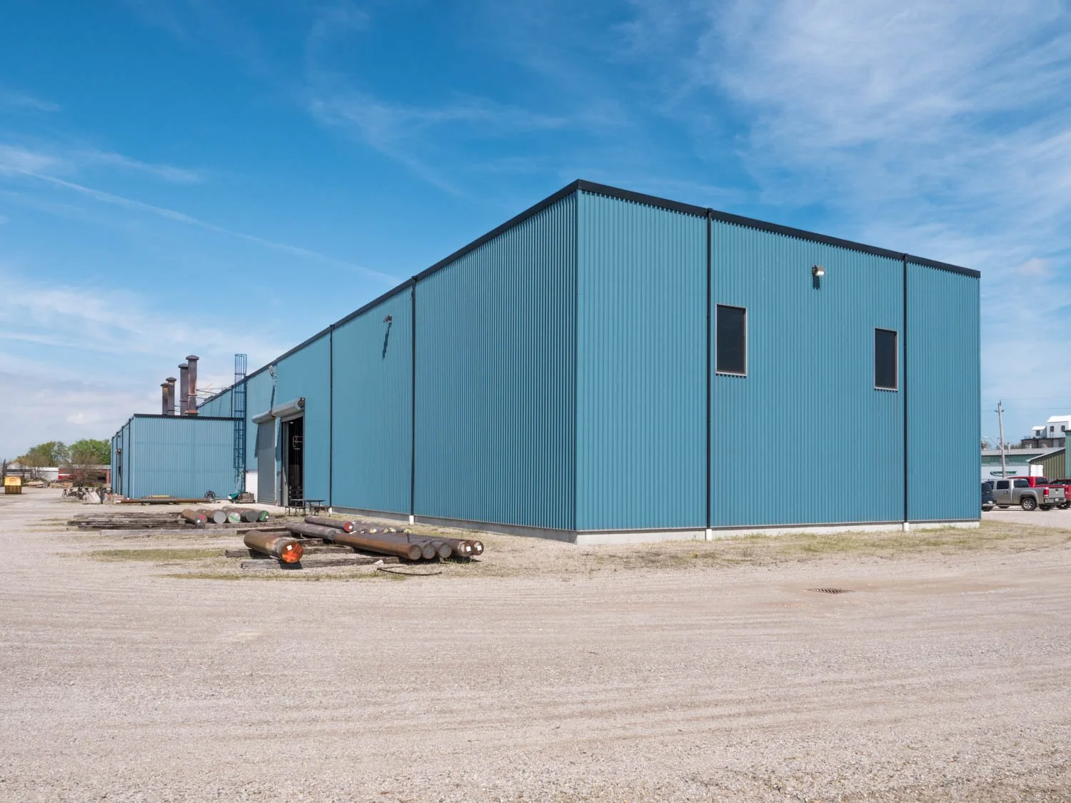 Blue industrial building with metal siding, small windows, and an outdoor area with pipes and gravel ground under a partly cloudy sky.