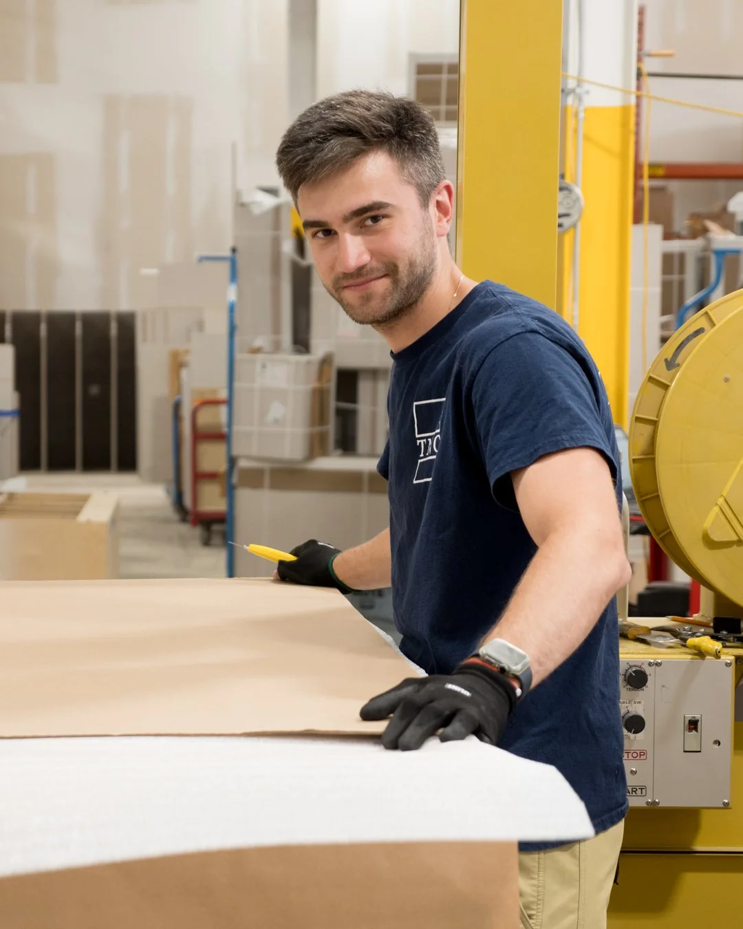 A man working in a workshop, handling a large sheet of material, wearing gloves, and smiling at the camera.
