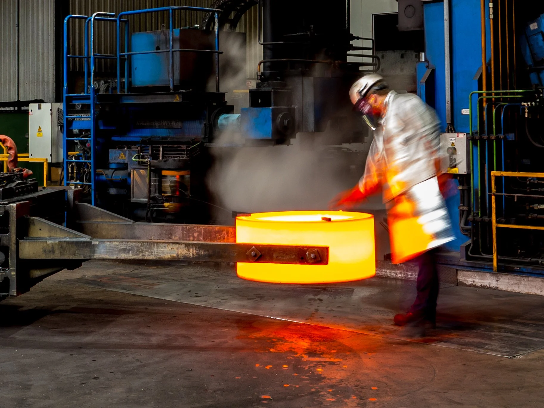 A worker in safety gear, including a helmet, glasses, and protective clothing, is working in a metal foundry with molten metal pouring out from a large furnace or container, creating bright orange sparks and steam.