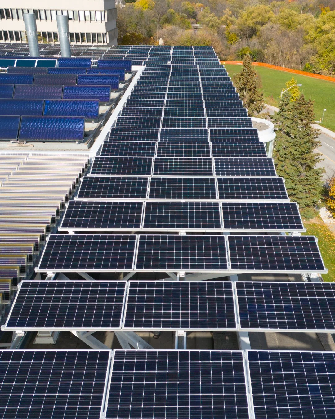 A large array of solar panels installed on the rooftop of a building, with some trees and a green field with orange fencing in the background.