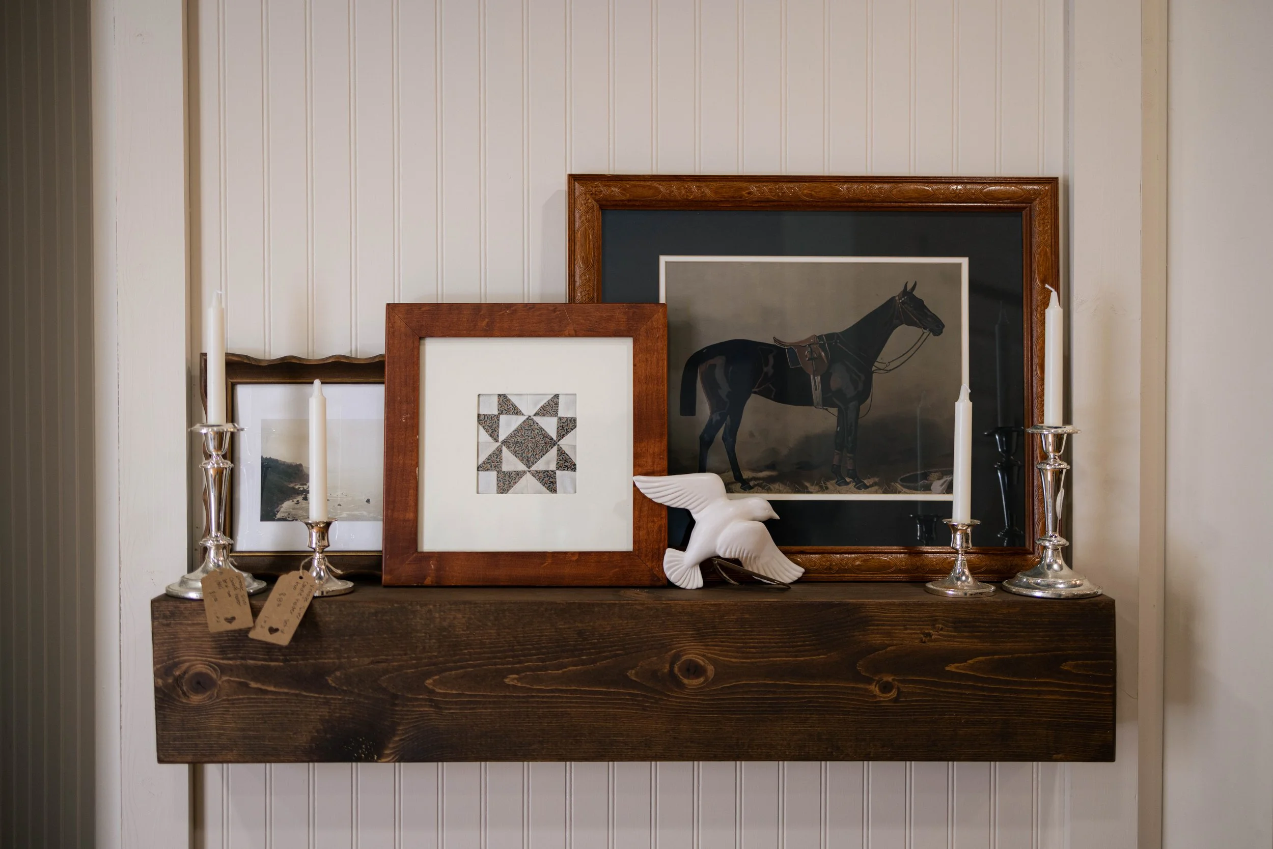 A display of framed artwork on a wooden shelf, with two tall silver candlesticks and a white sculptural figure of a reclining horse in front. The background is a white paneled wall.