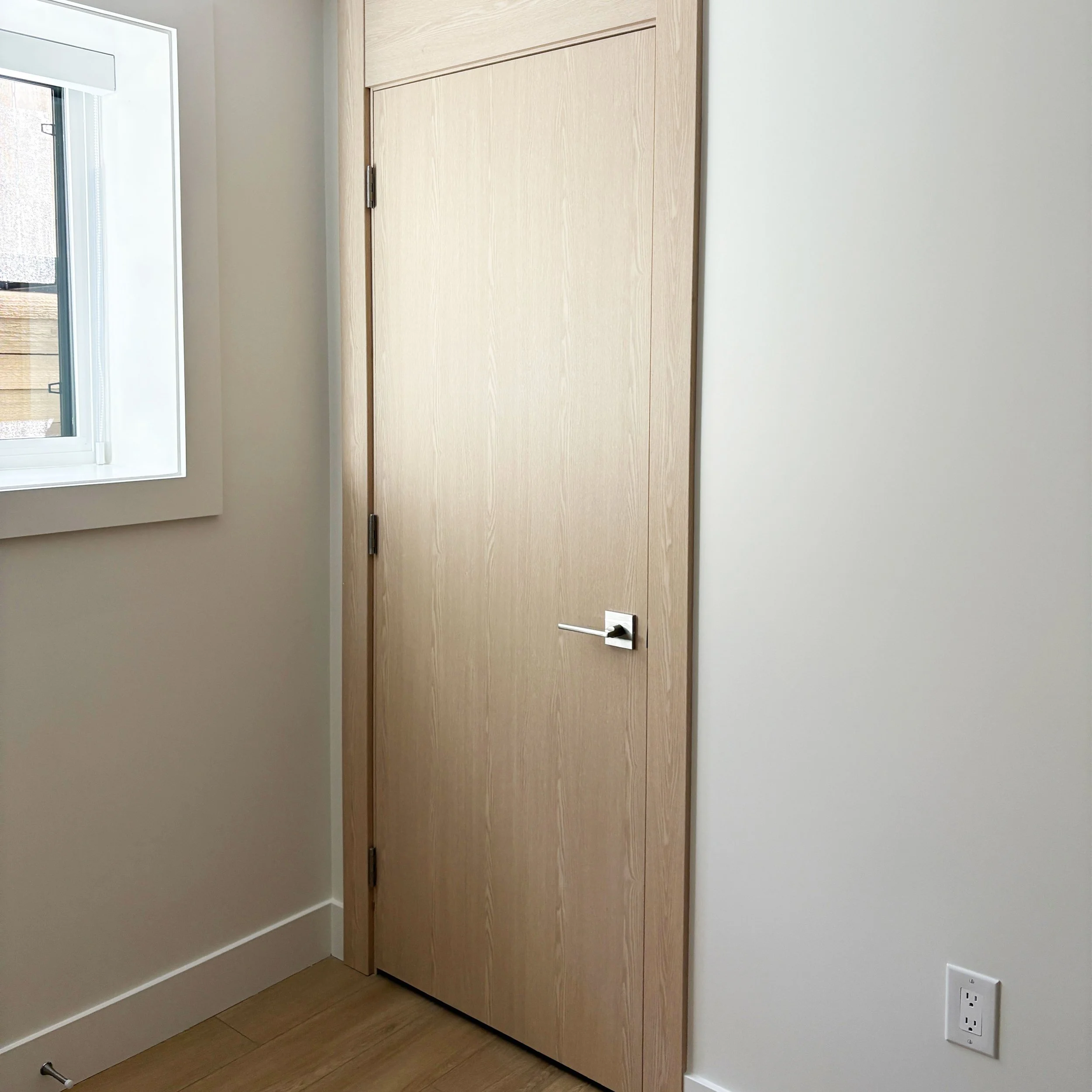 Interior room with a closed wooden door with a metallic handle, a window to the left, and an electrical outlet near the bottom right corner.
