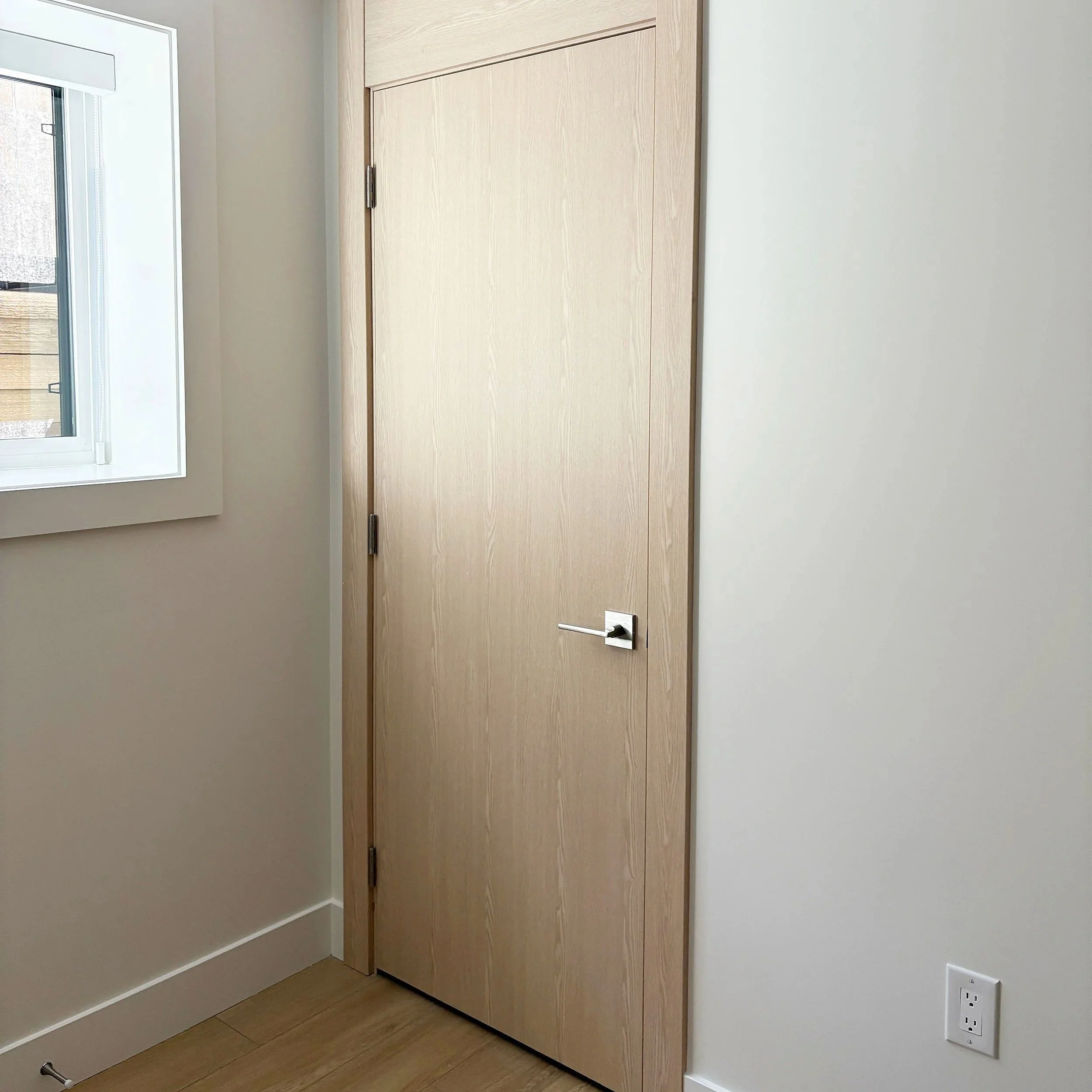 A closed wooden door with a modern silver handle in a room with white walls, a window, and a white electrical outlet.