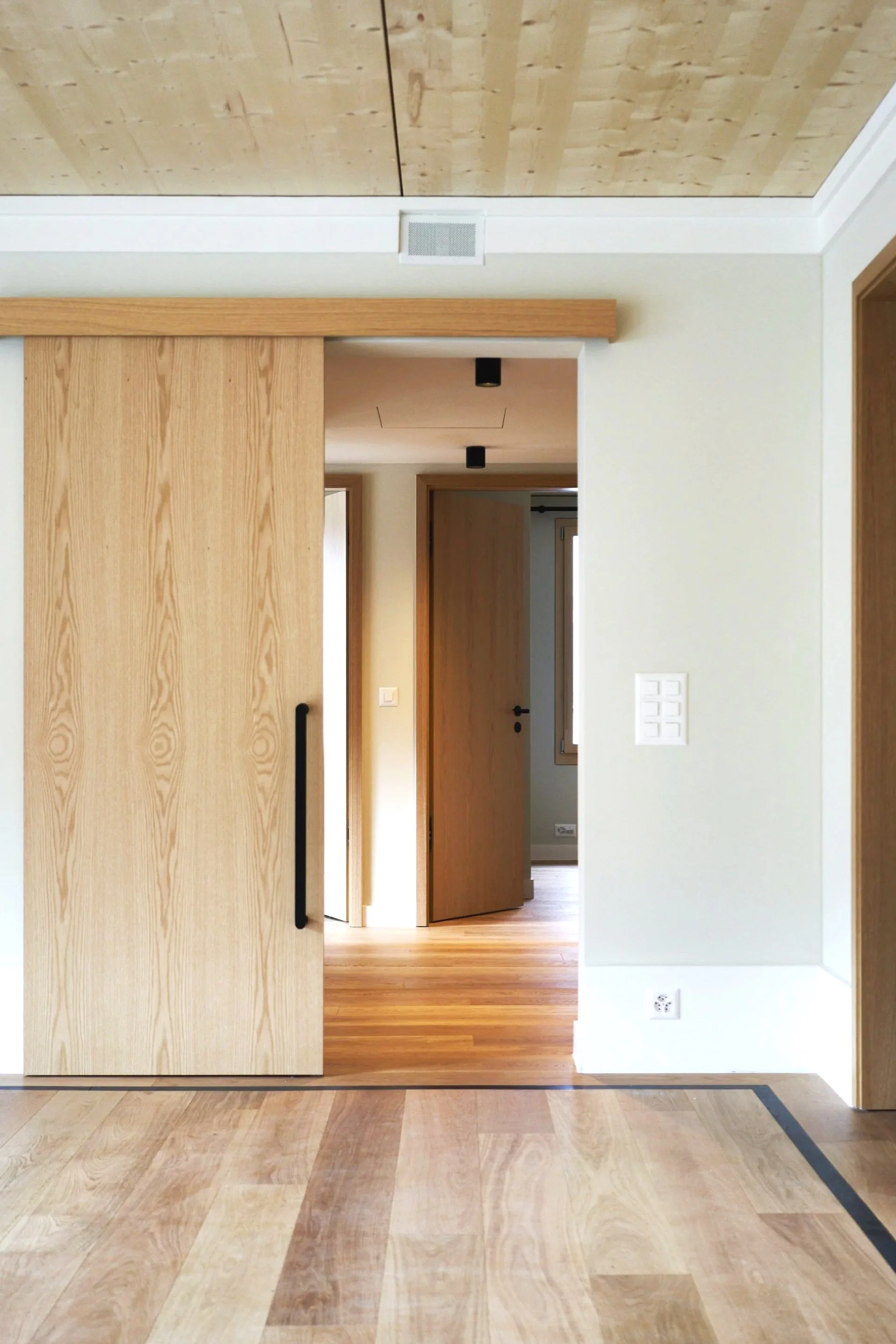 Interior view of a modern home with light wood doors, wood flooring, white walls, and ceiling with wood paneling.