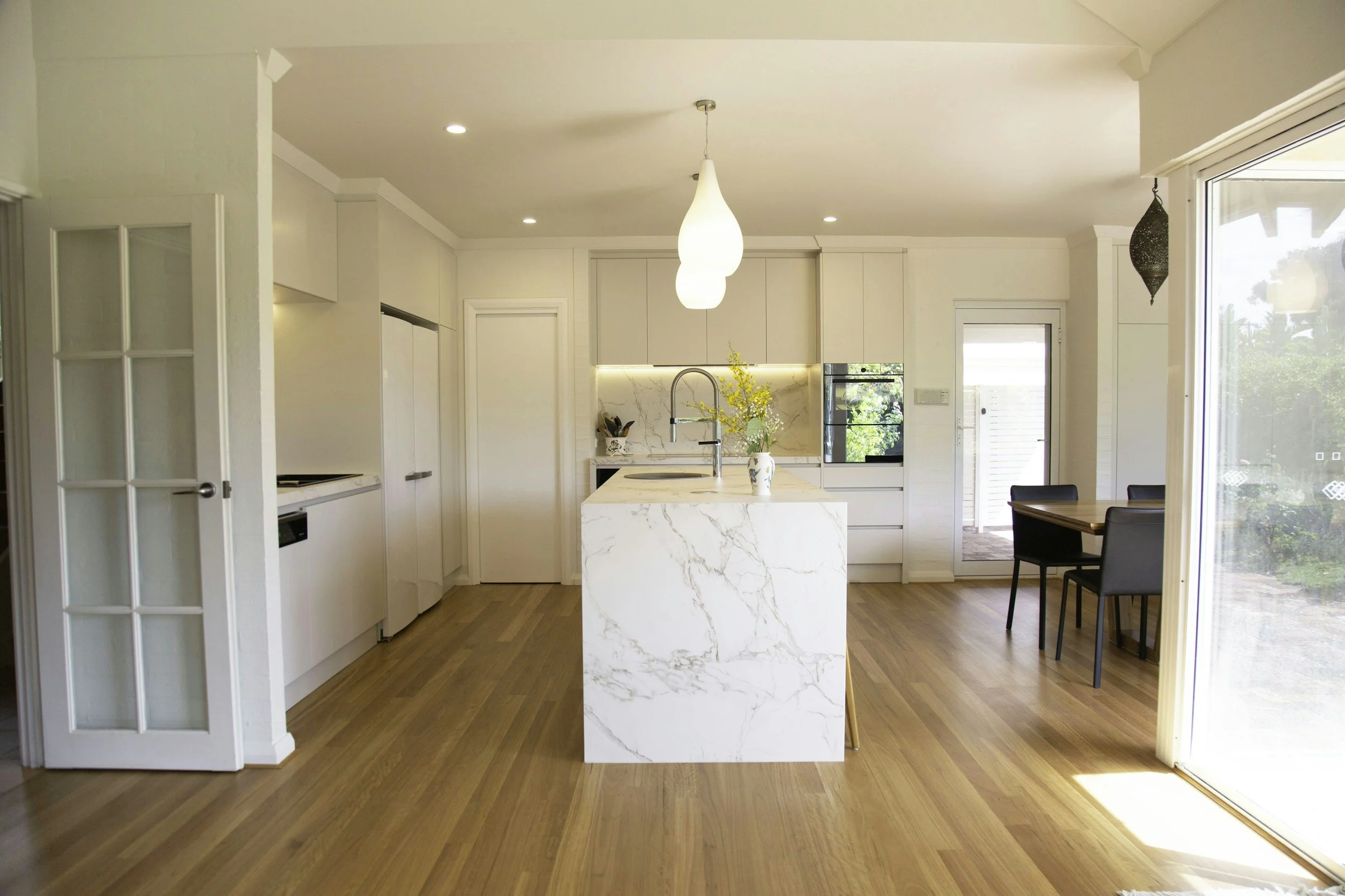 Modern kitchen with white cabinetry, marble island, warm colored wooden floors, and a dining area with black chairs. Natural light streaming in through large sliding glass doors. Warm Toned Flooring.