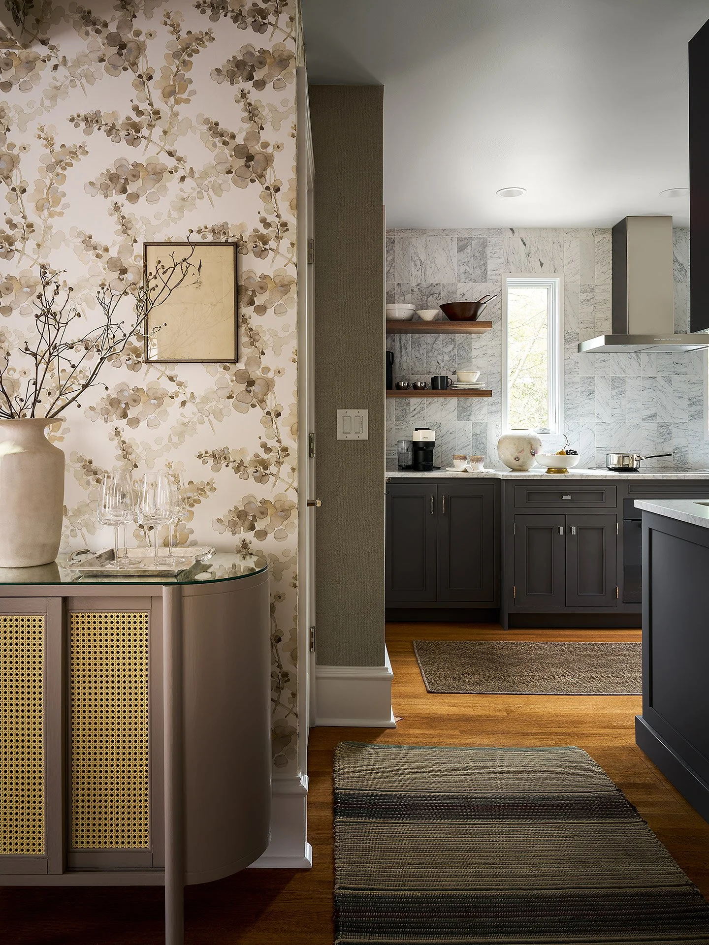 A kitchen with gray cabinets, white marble backsplash, and a small window, visible from an adjacent room with floral wallpaper and a sideboard with glassware and a vase.