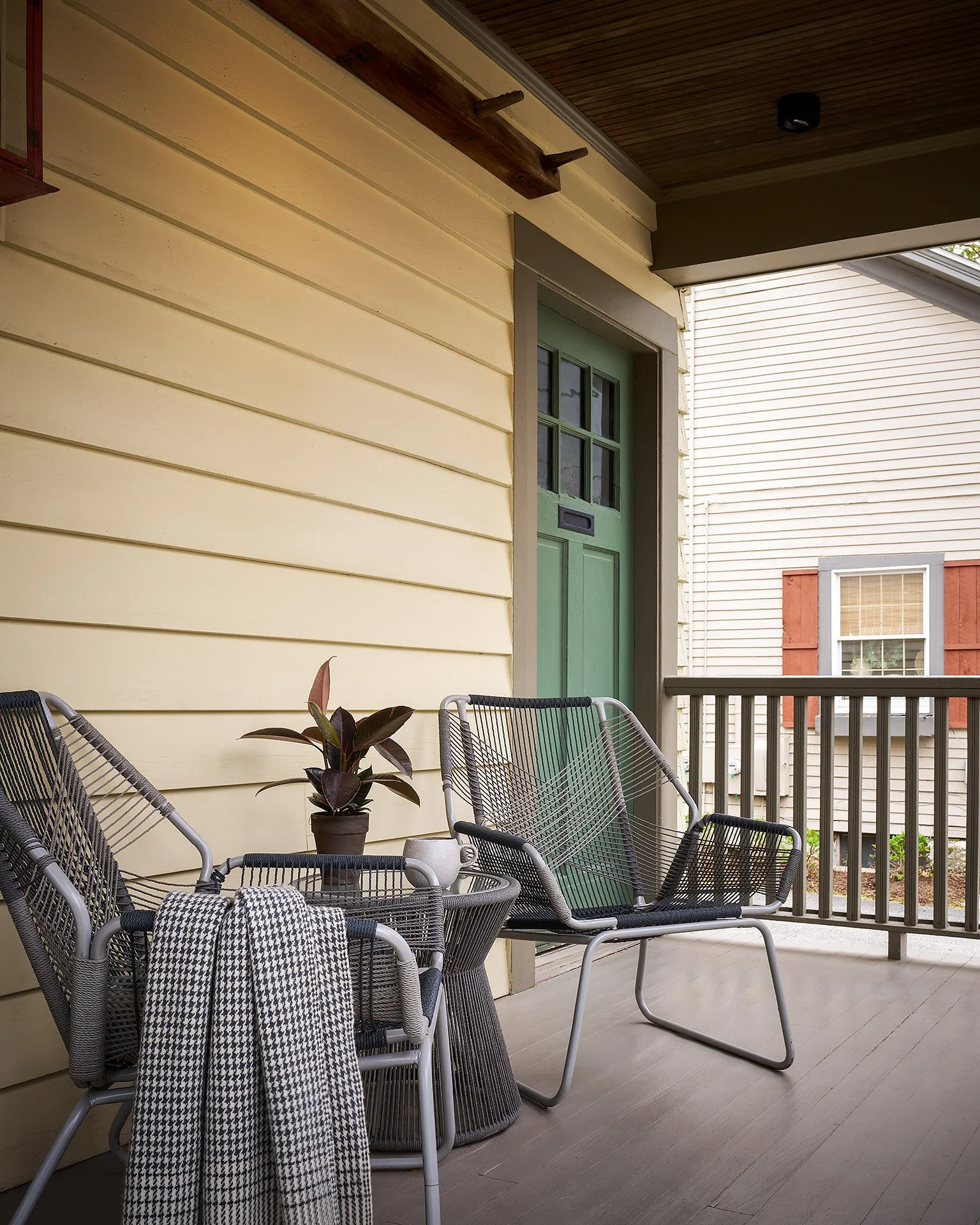 Front porch with yellow siding, green front door with mail slot, black patio furniture, potted plant, and railing, neighboring house with window and shutters visible in the background.