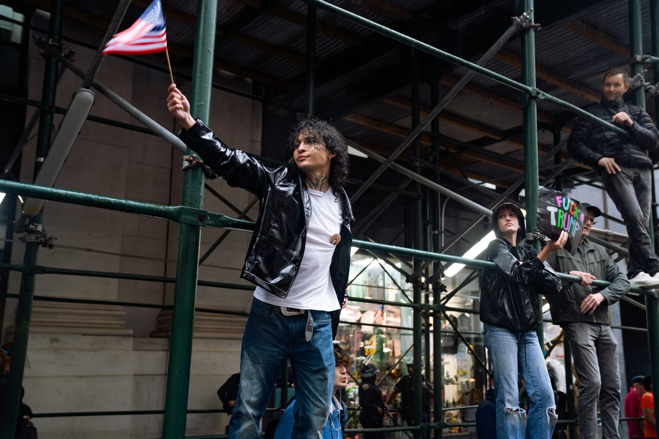 Protesters standing on scaffolding, one woman waving an American flag, another woman holding a sign that says 'F*** Trump', in a city street.