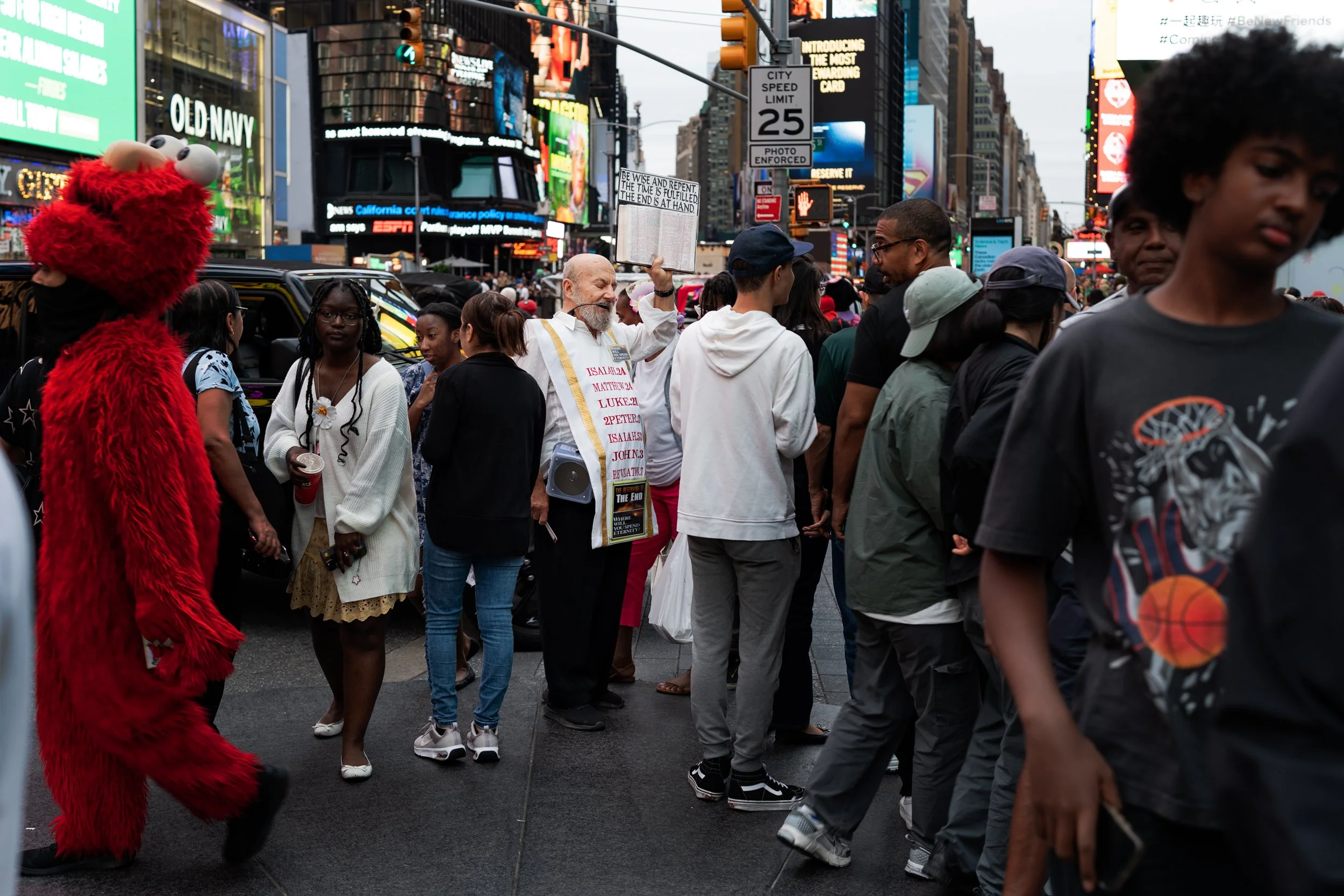 People standing in line in Times Square, New York City, with bright billboards and digital screens in the background. A person dressed as Elmo from Sesame Street is on the left side of the image. A man holding up a sign with written text is in the ce
