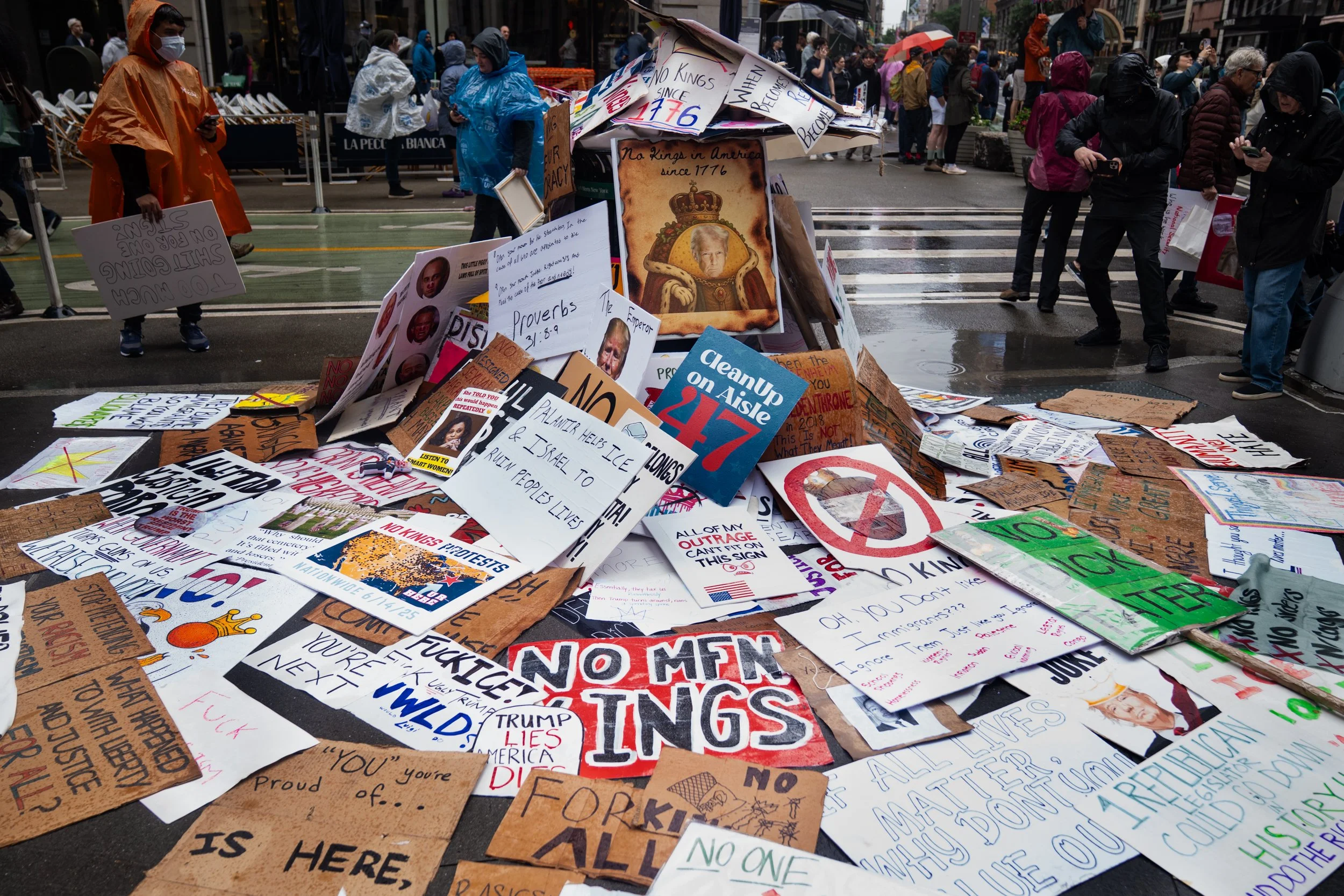 Protest signs and posters on the street with people holding signs, some wearing masks and rain gear, during a protest against a perceived threat from non-American entities and advocating for social justice.