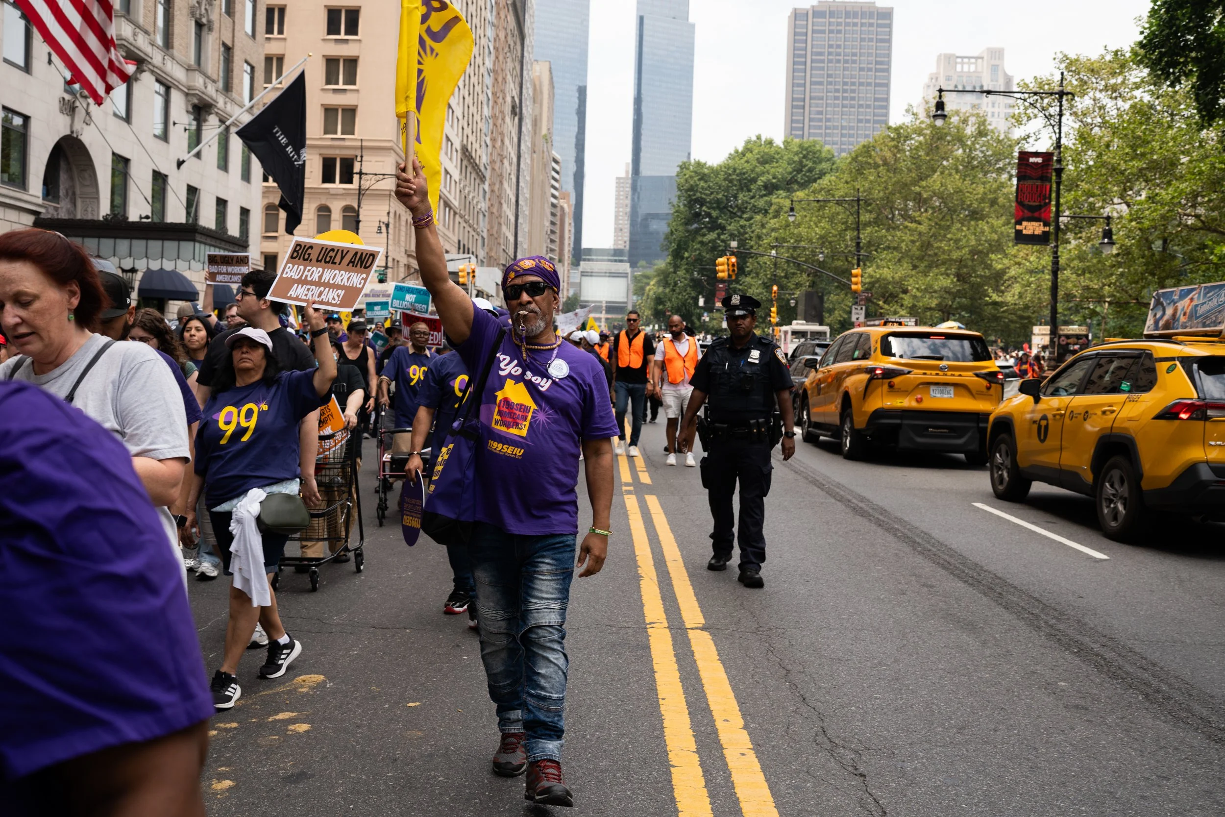 A protest march on a city street with numerous participants, some holding signs and flags, with police officers nearby and yellow taxis on the side.