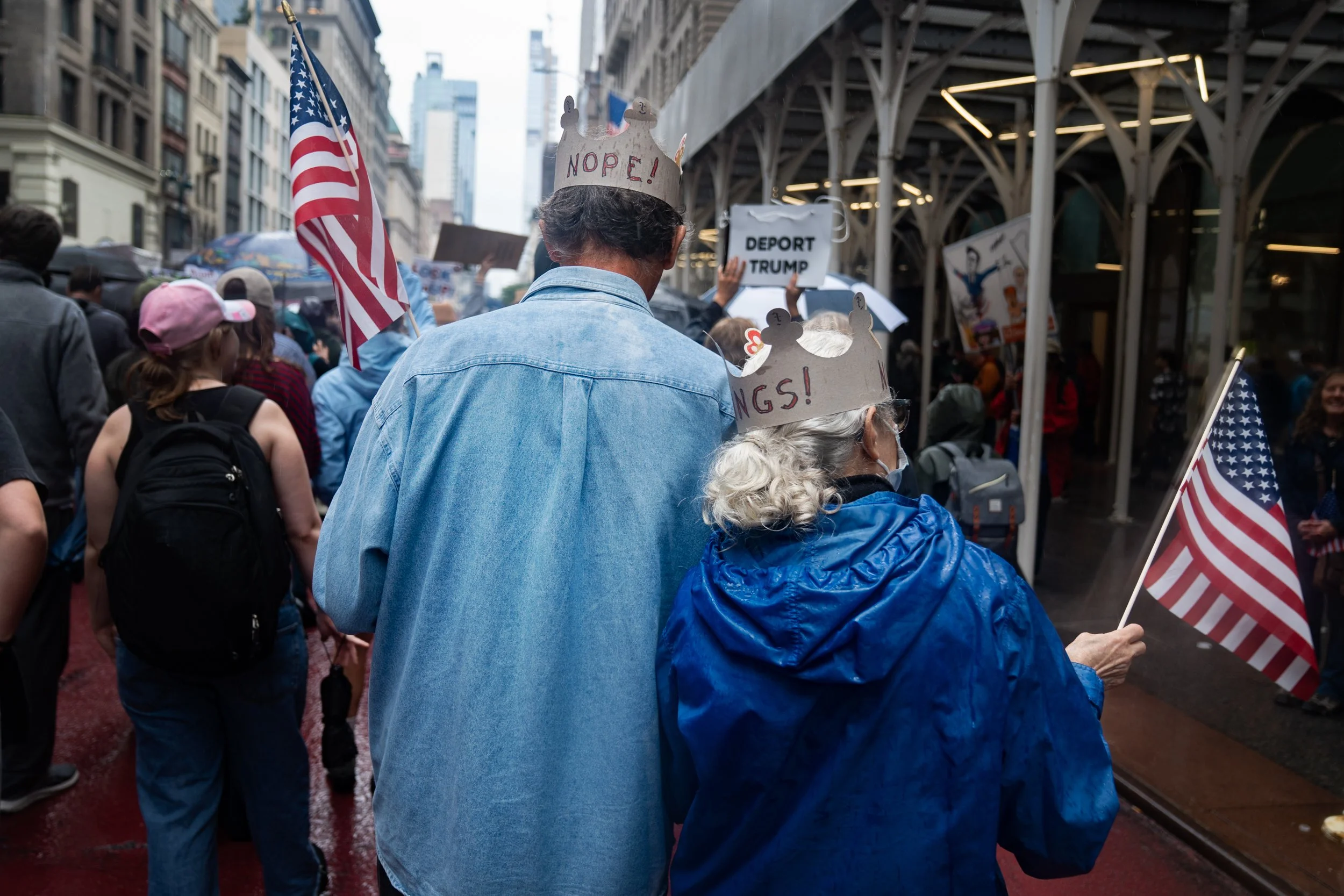 People participating in a protest or rally, many wearing crowns labeled 'NOPE!' or 'WINGS!', holding American flags, with signs that include 'DEPORT TRUMP', on a city street under a covered area.