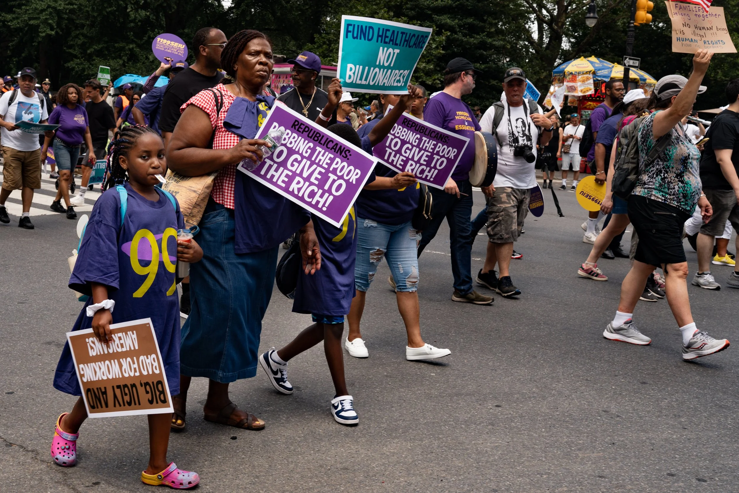 Protesters marching with signs advocating for healthcare funding and addressing economic inequality, including a woman and a girl holding signs on a city street.