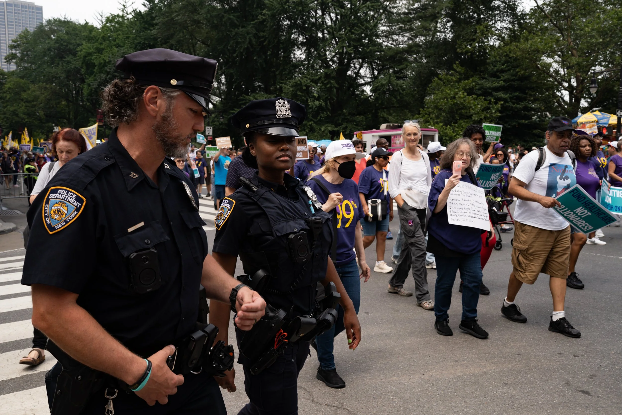 Police officers and protesters marching in a demonstration, holding signs and banners, with a crowd and trees in the background.