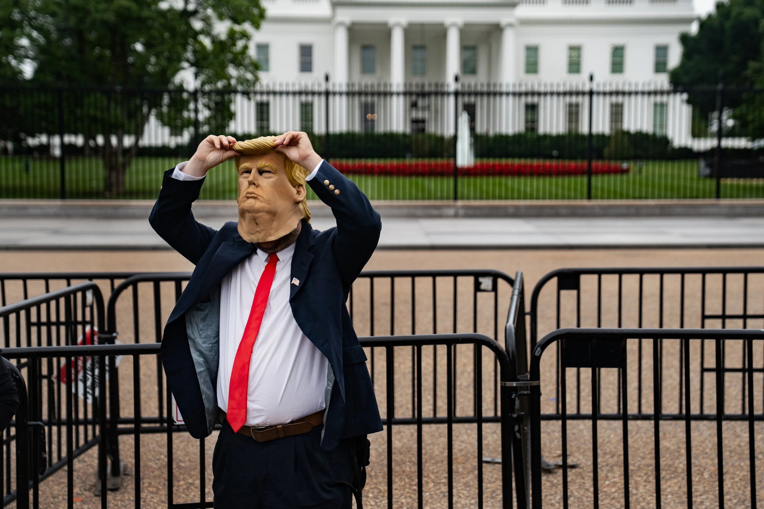 Person wearing a Trump mask, dressed in a suit with a red tie, standing outside in front of the White House with barriers around, appearing to adjust the mask.