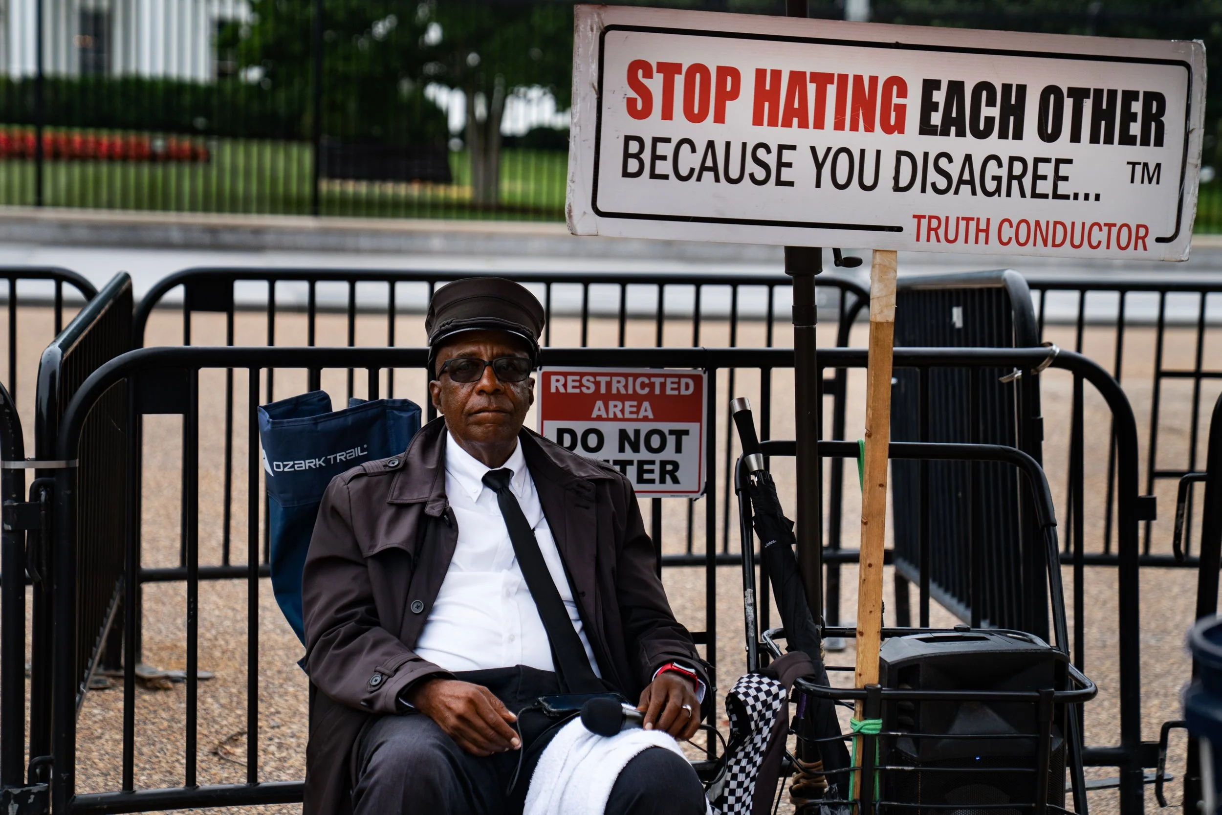 A man sitting behind a fence with two signs, one reading 'Stop Hating Each Other Because You Disagree,' and another 'Restricted Area Do Not Enter.'
