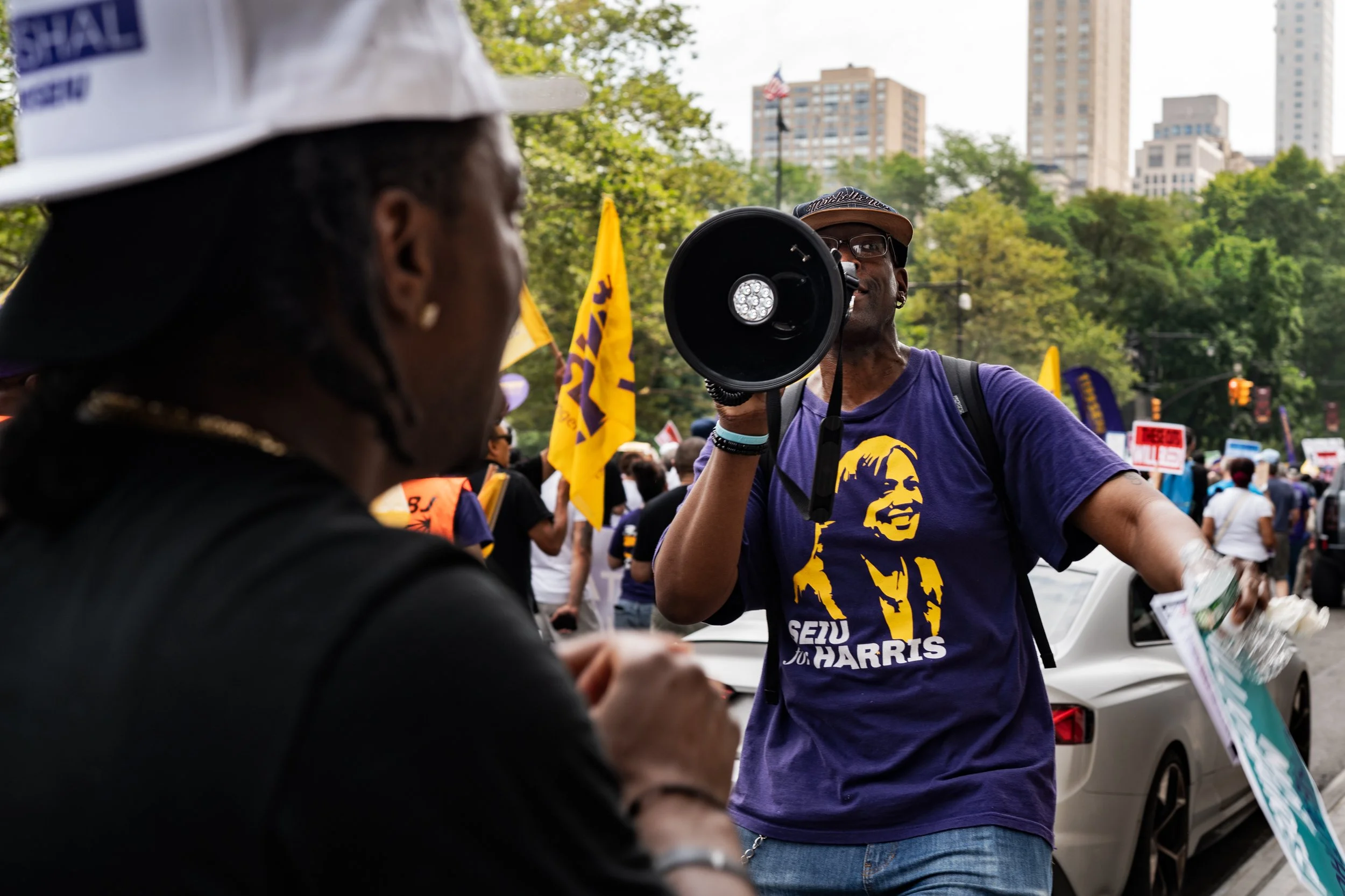 A man wearing a purple 'Seize the Moment to Harris' T-shirt and holding a megaphone speaking to another person at a march or rally in a city park with tall buildings in the background.