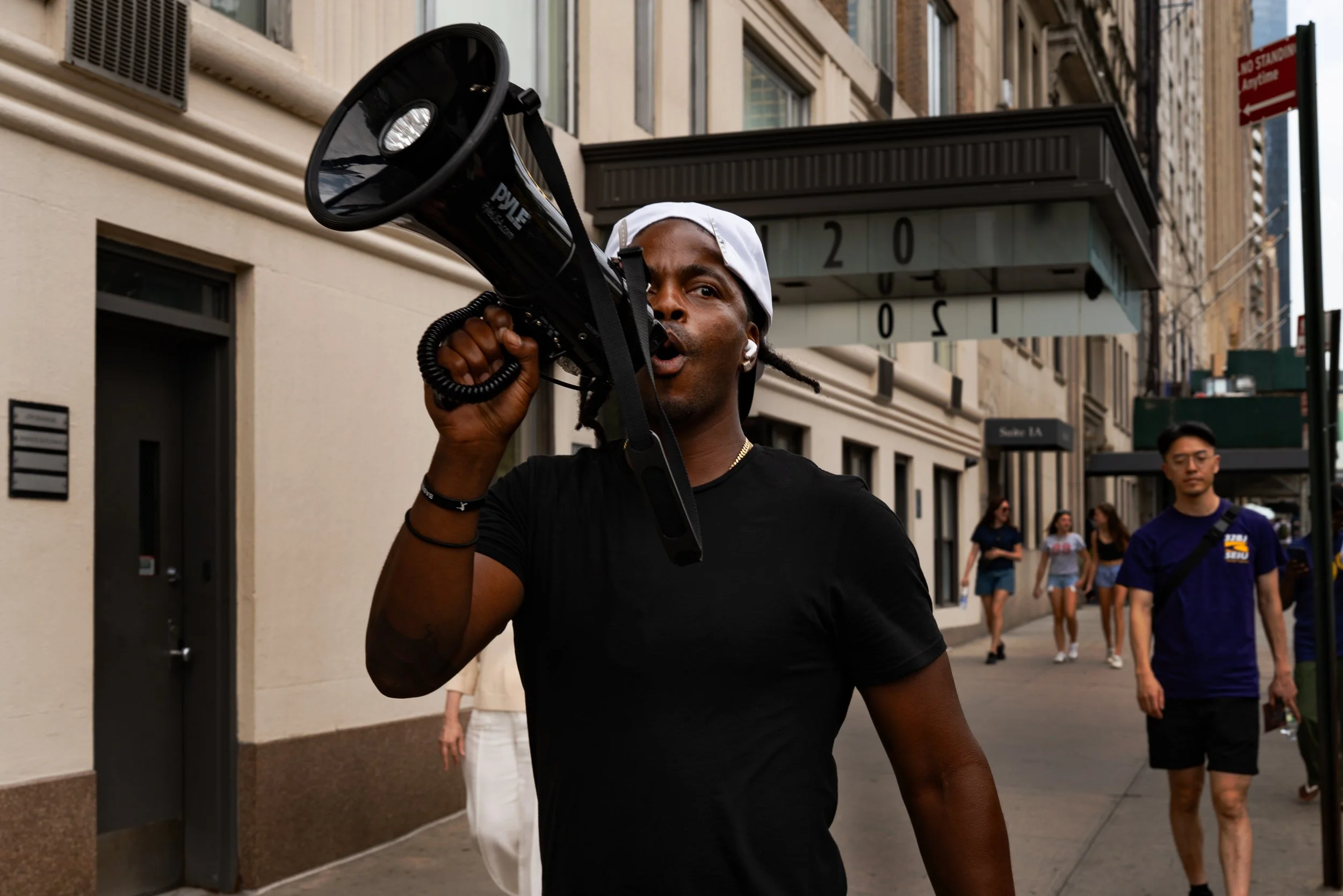 Man speaking into a megaphone on city sidewalk, with pedestrians in the background.