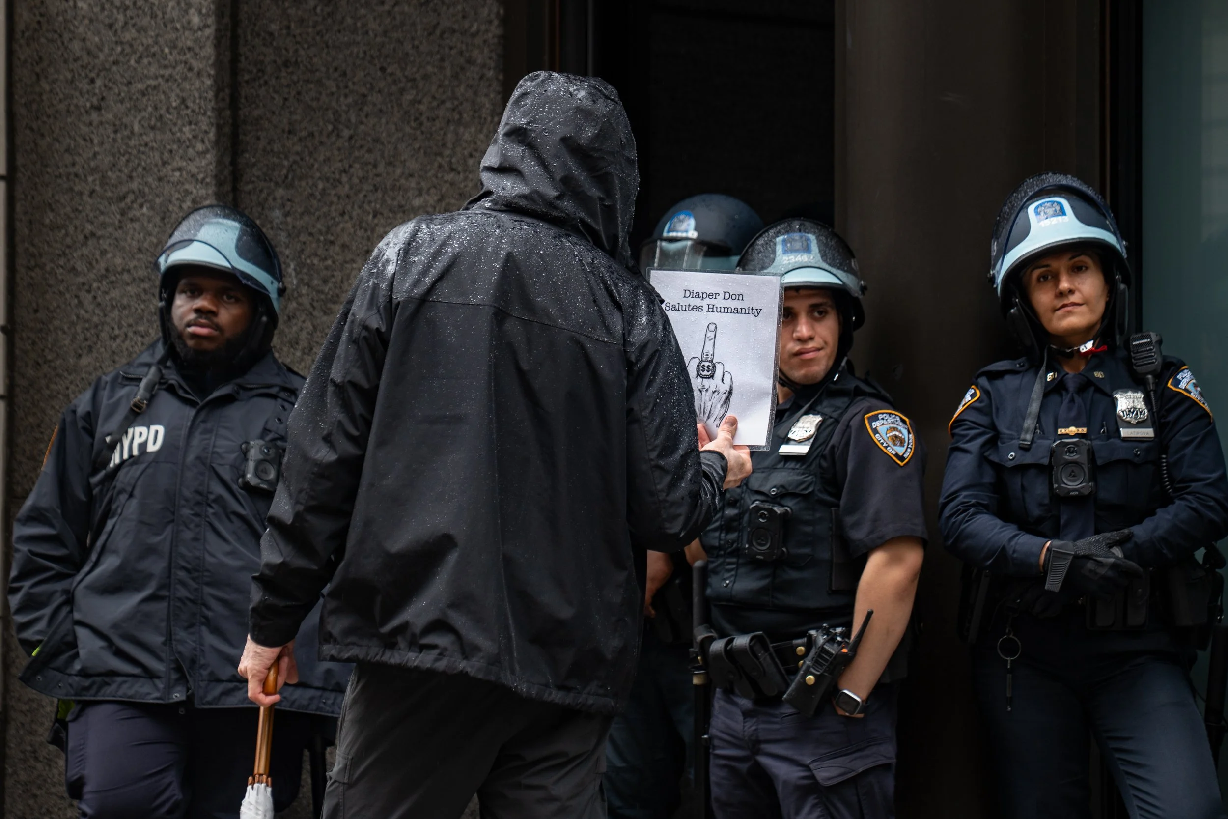 A man holding a sign with a hand showing the middle finger and the words 'Diaper Don Salutes Humanity' is confronting three police officers in uniform during a rainy day.