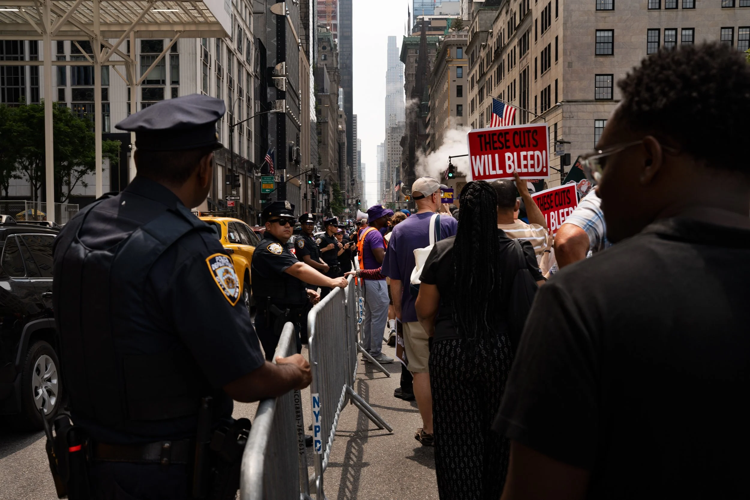 Protest on a city street with police officers and demonstrators holding signs that read 'These cuts will bleed!' amidst tall buildings.