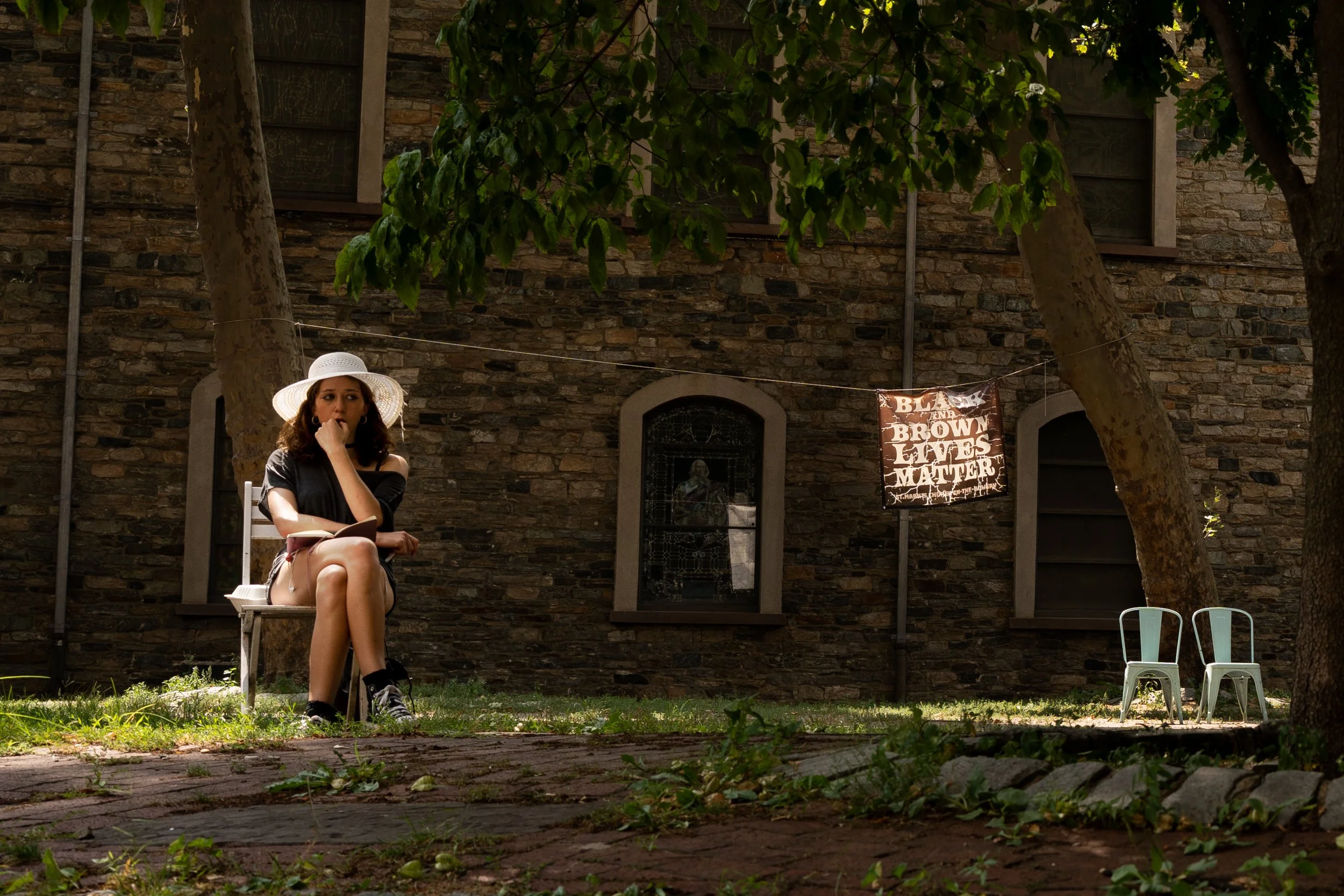 A woman sitting on a white chair under a tree in front of a brick building, wearing a wide-brimmed hat and a black dress, with two small white chairs beside her and a sign that reads 'Black Lives Matter' hanging on a wire.