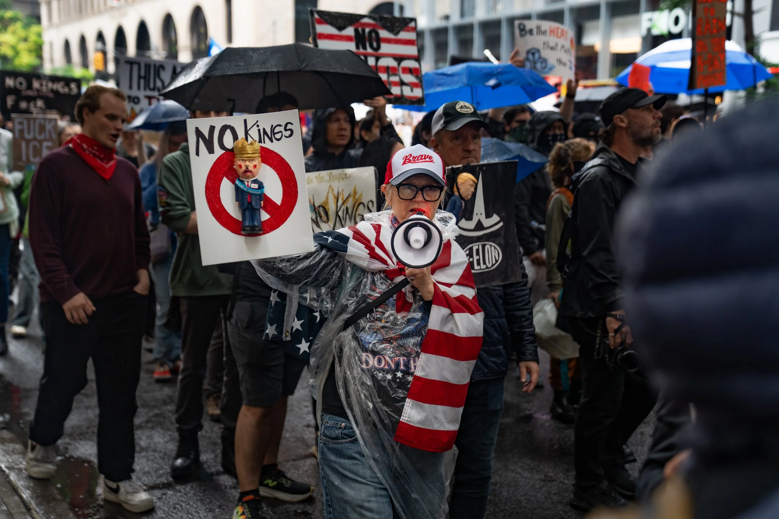 Protesters marching in the rain holding signs and umbrellas, some wearing patriotic and political clothing, with a woman in the foreground speaking into a megaphone, wrapped in an American flag, during a demonstration.