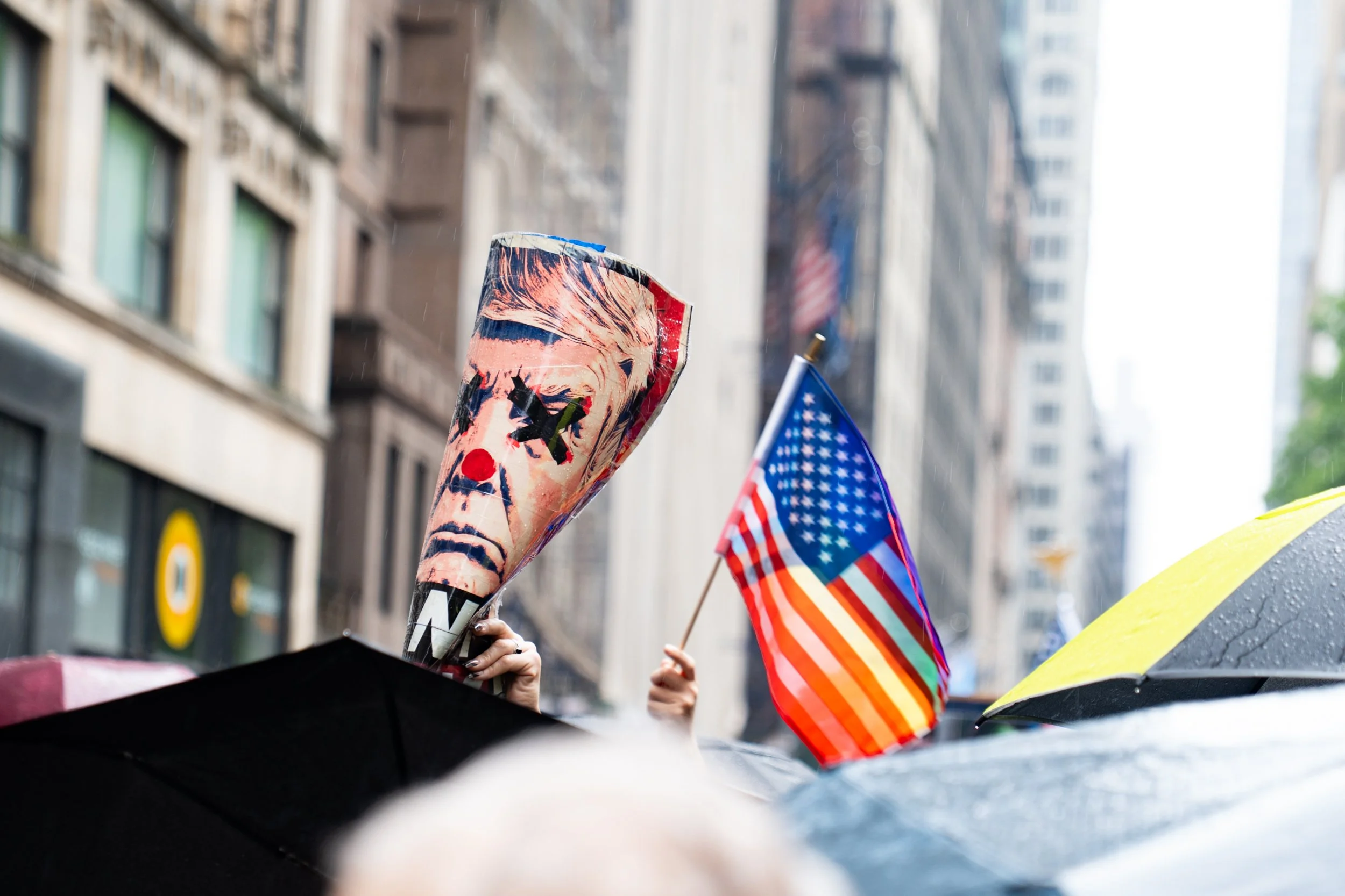 Crowd holding protest signs and flags, including an American flag and a Trump-themed sign, on a rainy city street with tall buildings.