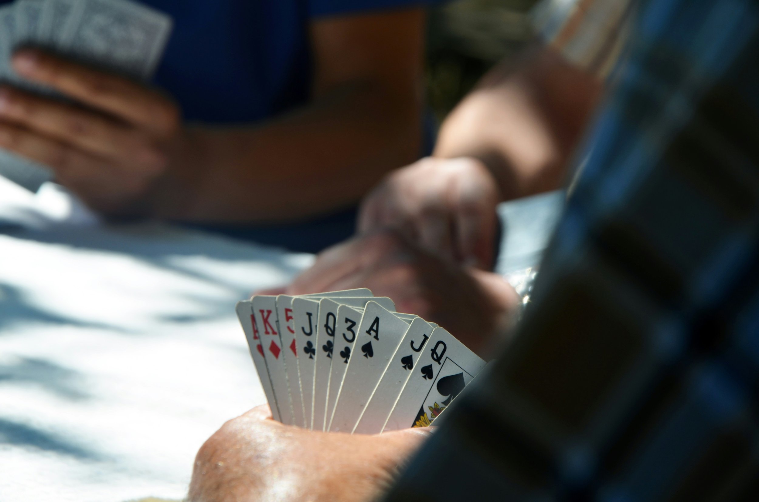 Close-up of a person holding a fan of playing cards, including king, queen, jack, and ace of spades, along with other cards, while sitting outdoors near a picnic table.