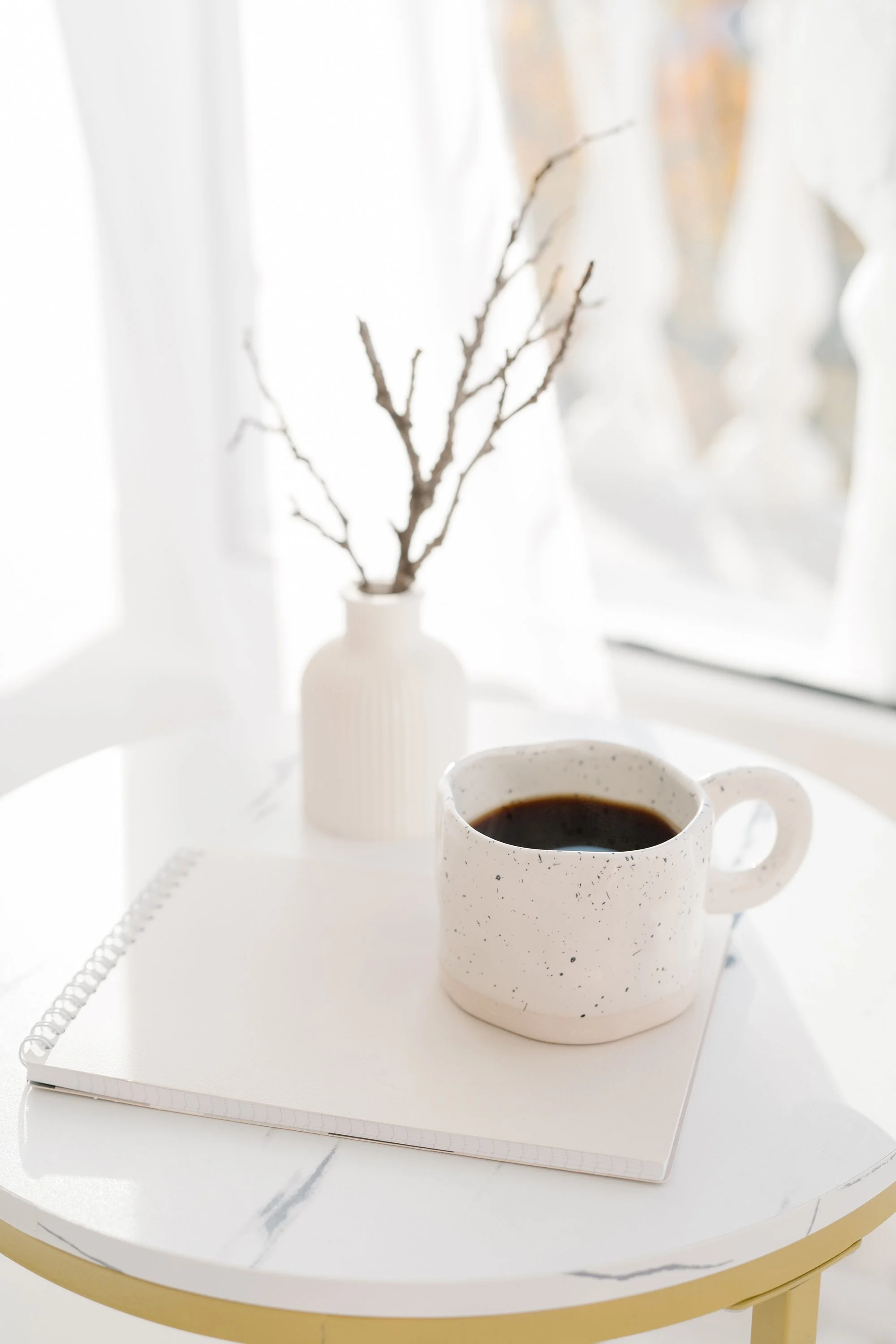 White ceramic mug with black coffee on a white notepad, on a white marble table next to a small white vase with bare branches, near a window with sheer white curtains.