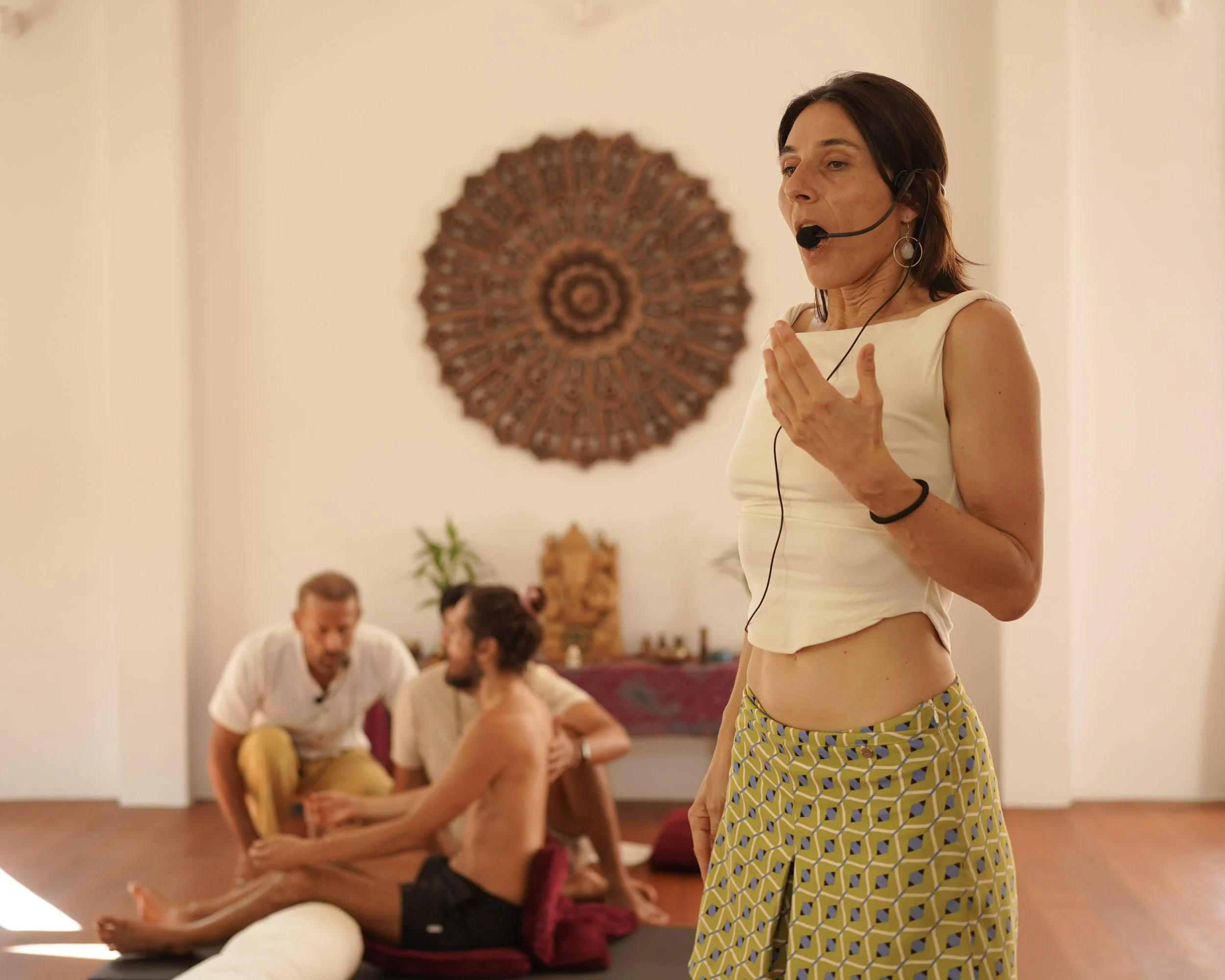 A woman with short dark hair, wearing a white sleeveless top and yellow geometric patterned pants, giving a spiritual talk with a microphone headset in a room with three men sitting on the floor in meditation or yoga pose.